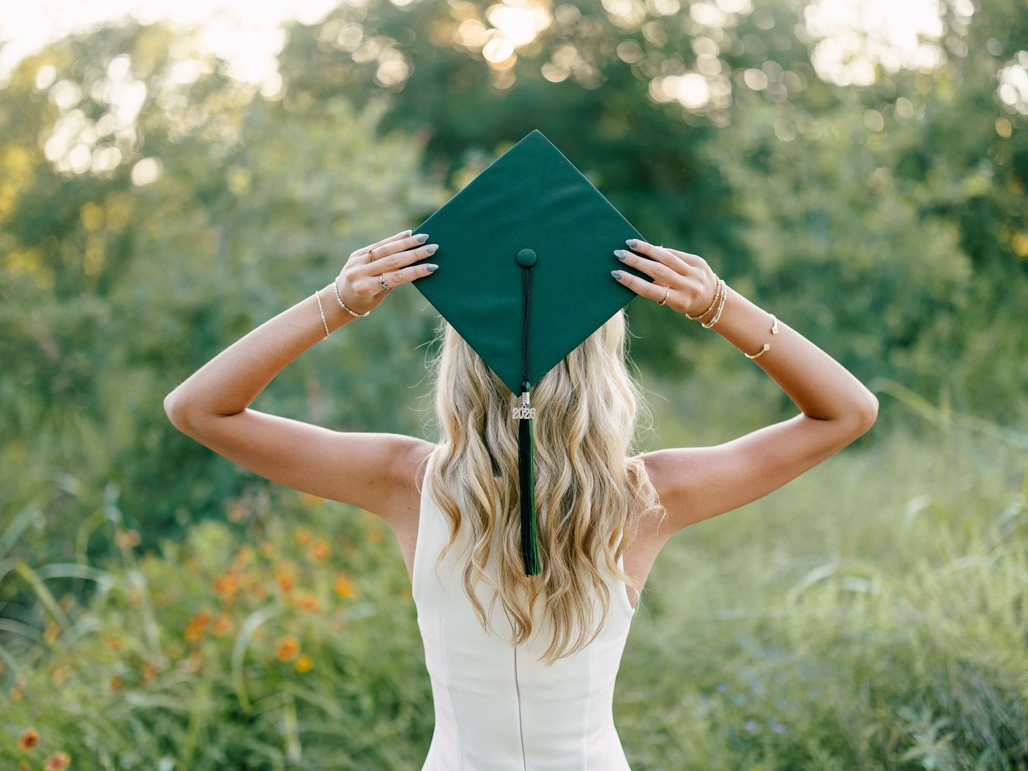 Young woman in white dress holding dark green graduation cap outdoors in lush green field for senior photos.
