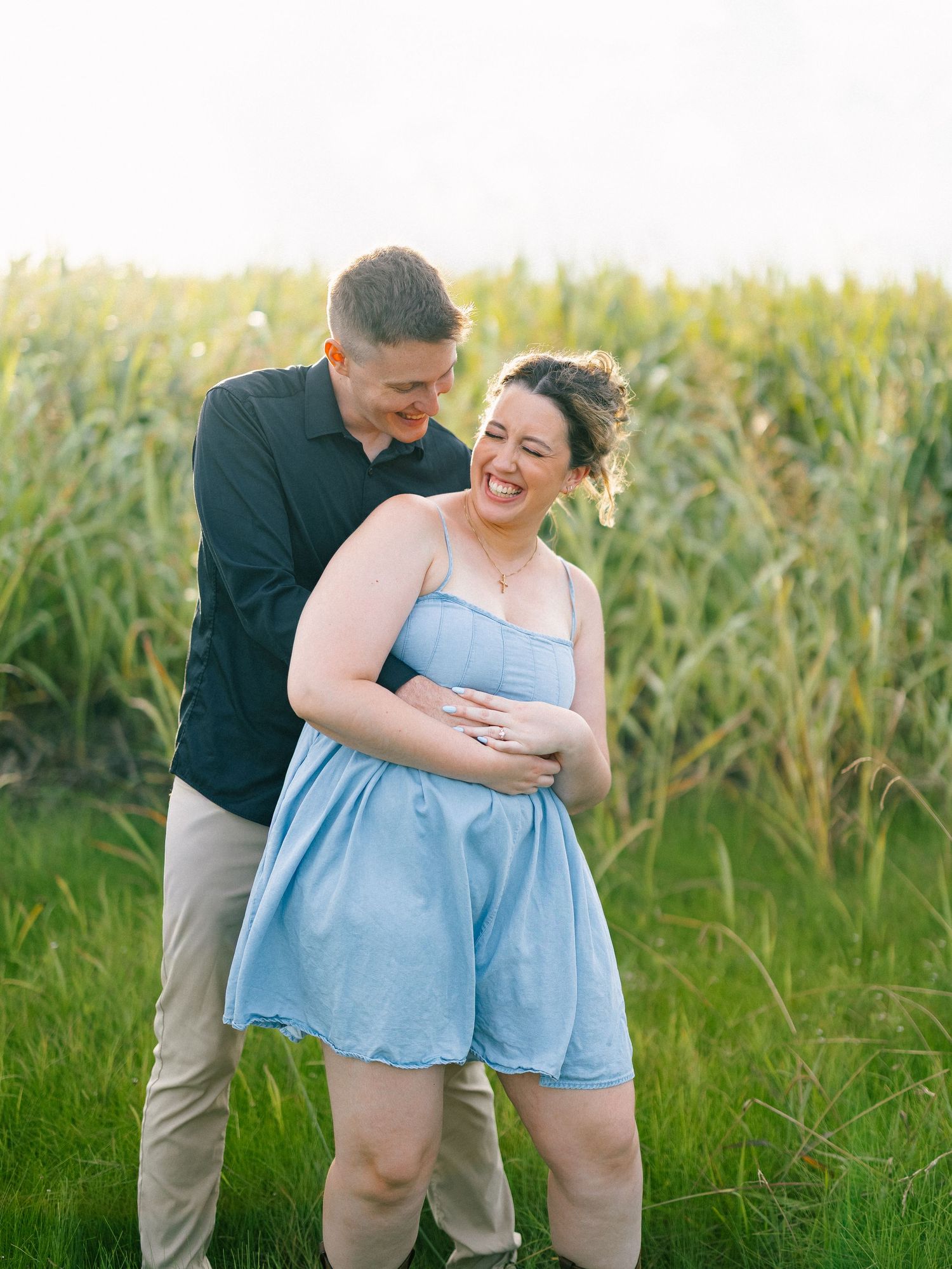 Couple laughing together in a green field, woman in blue dress embraced by man in black shirt during golden hour.