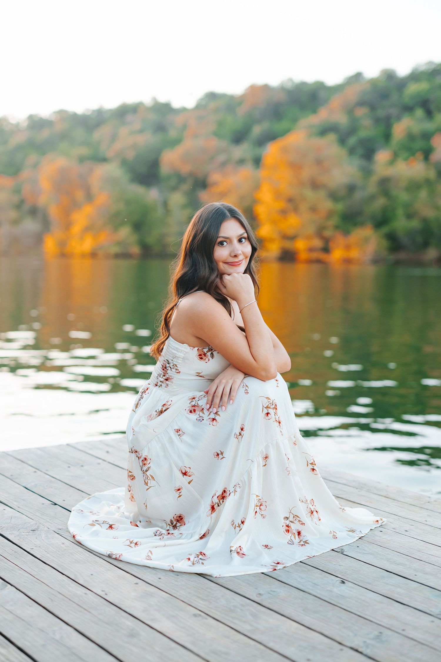 Young woman in floral maxi dress sitting on wooden dock by autumn lake with colorful foliage backdrop.