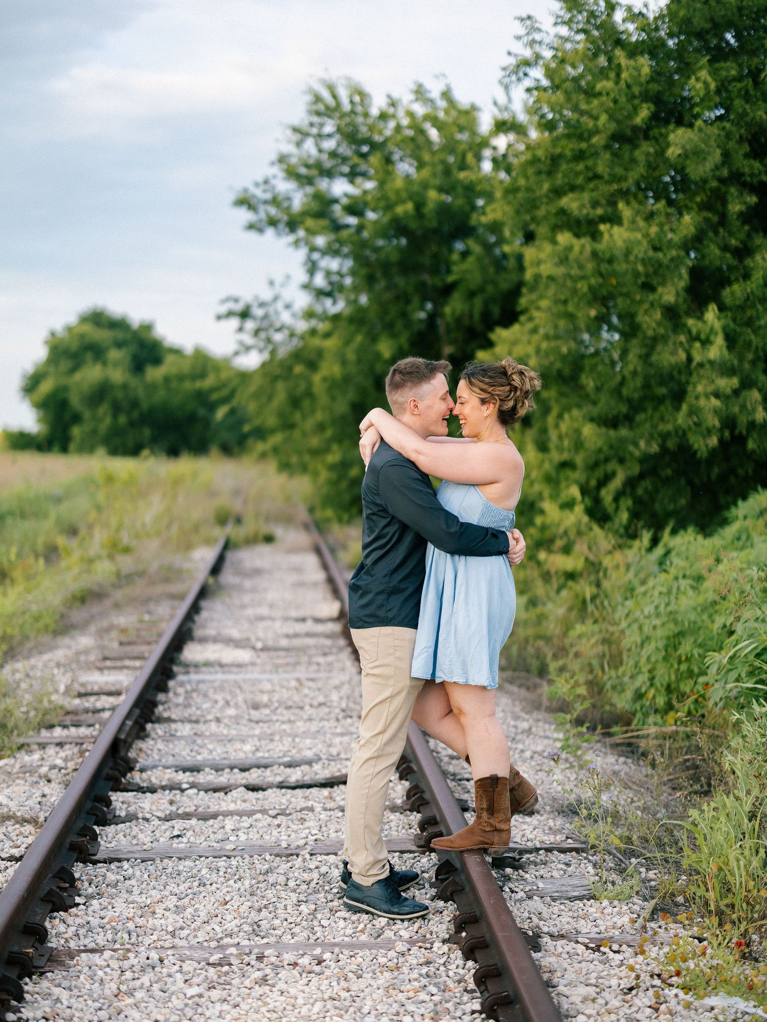 Couple embracing and almost kissing on railroad tracks surrounded by lush green trees during an outdoor engagement photo session.
