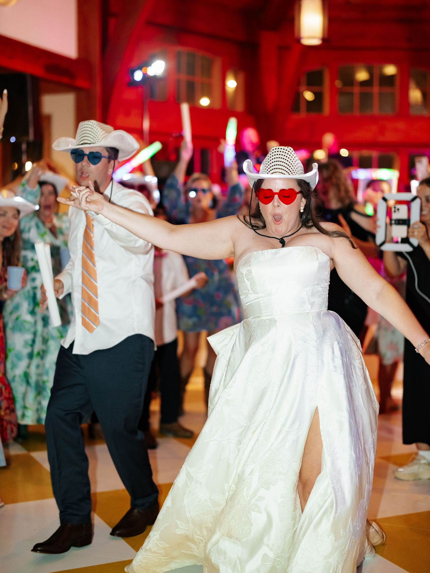 Bride and groom dancing energetically at wedding reception wearing fun accessories and cowboy hats.