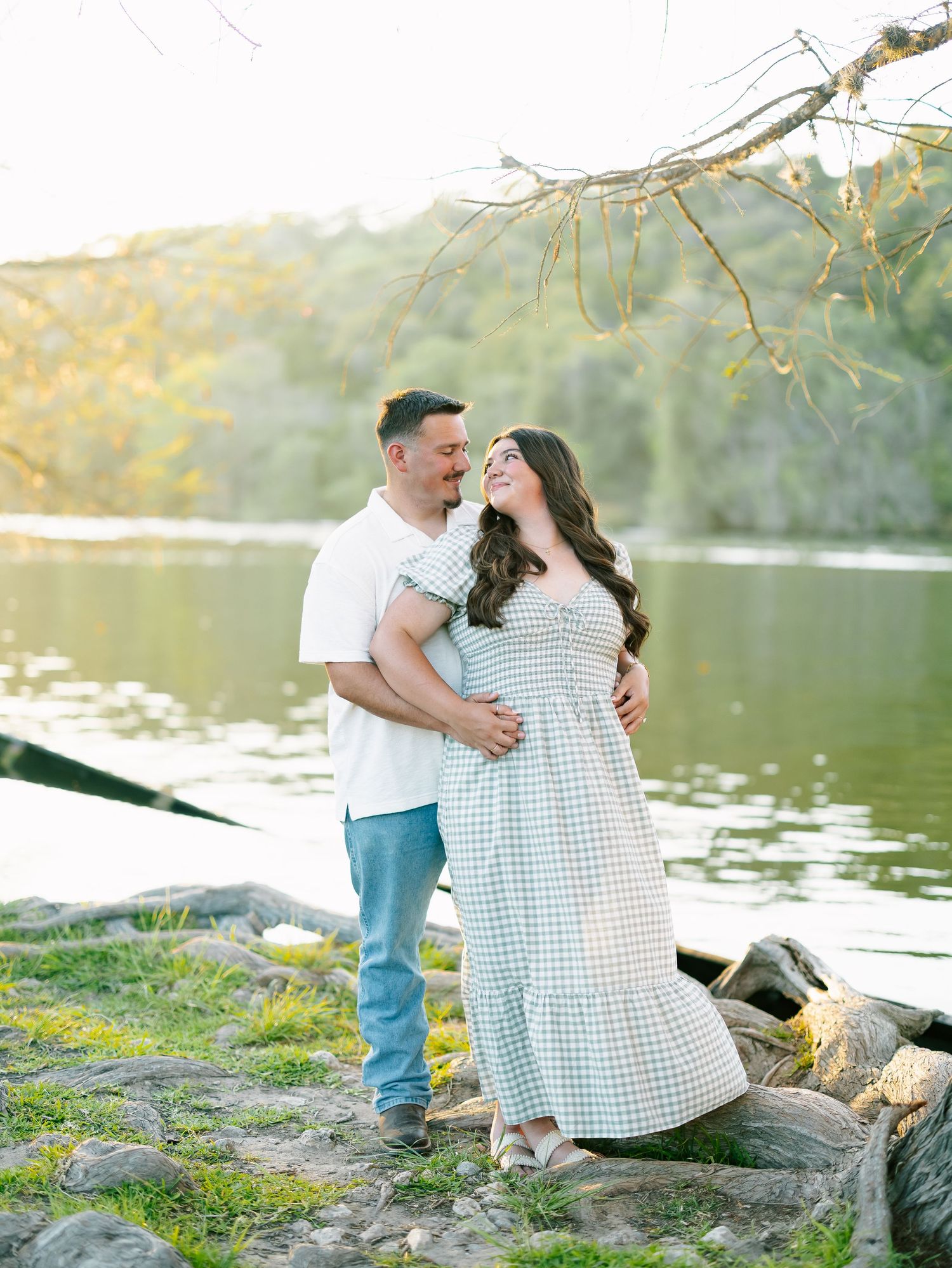 Engaged couple standing together by a peaceful lake surrounded by lush greenery at sunset.