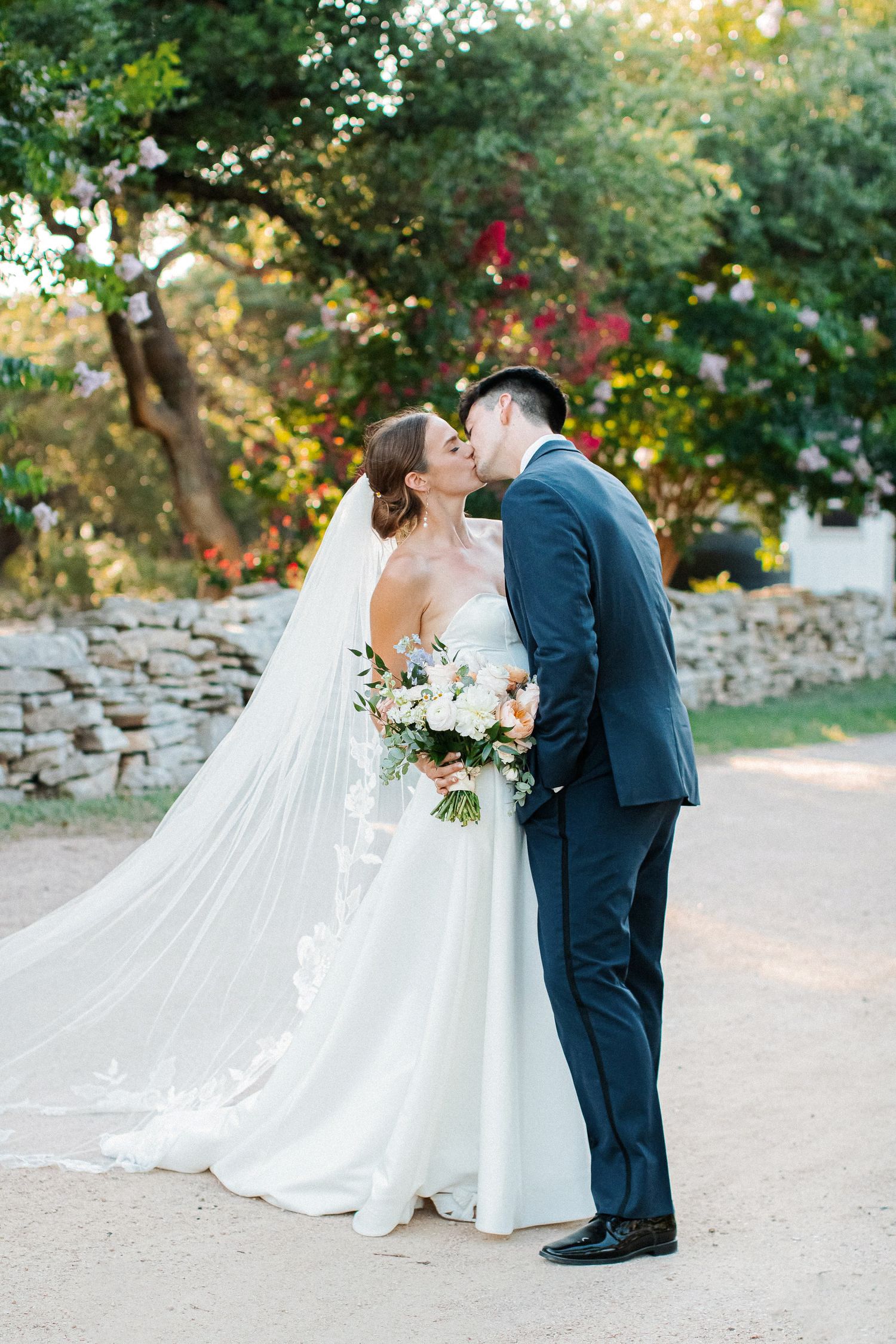 Bride and groom kiss outdoors with a flowing cathedral veil and colorful garden backdrop.