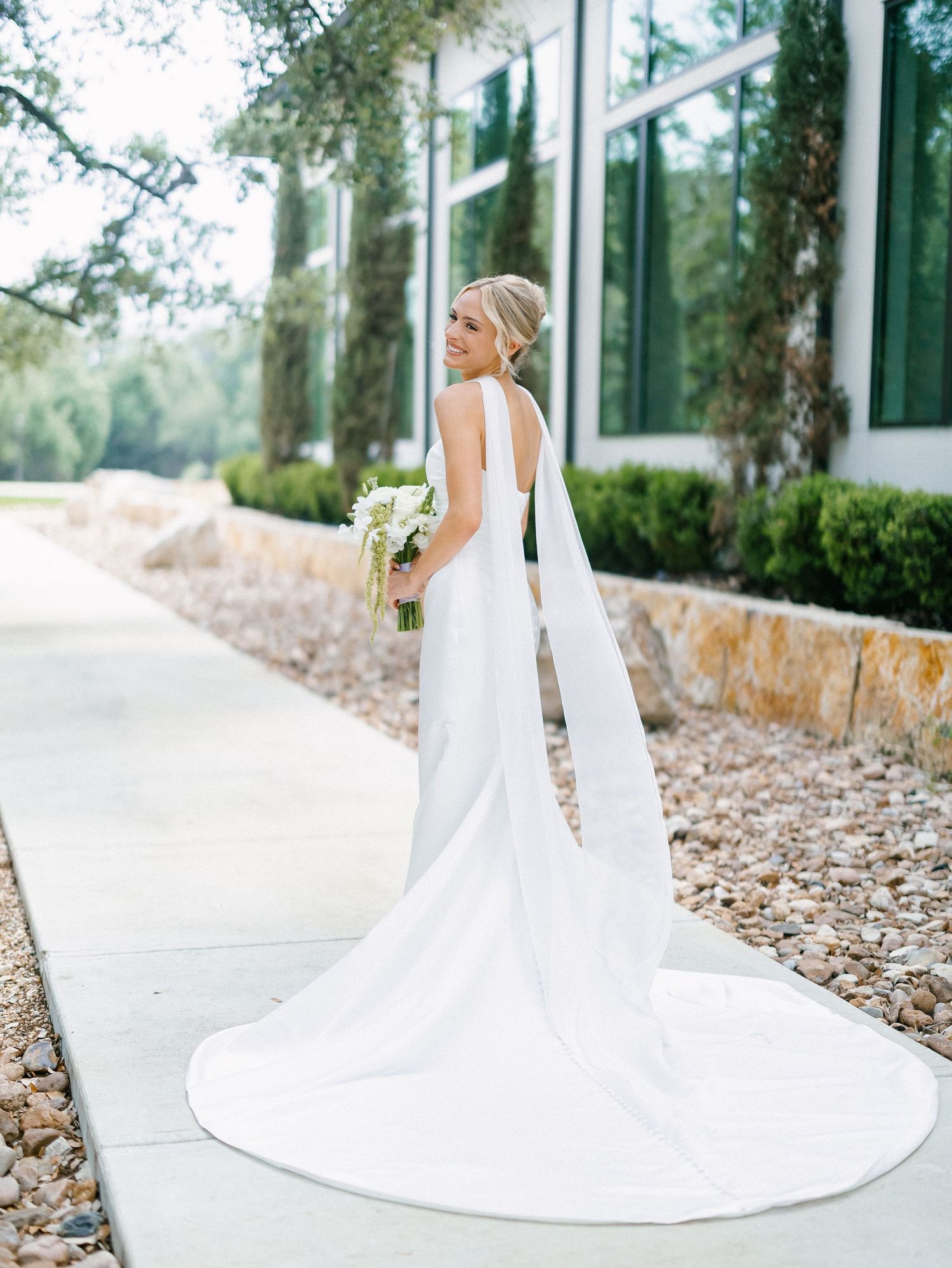 Elegant bride in a white halter gown with long train and veil holds bouquet outdoors near stone building.