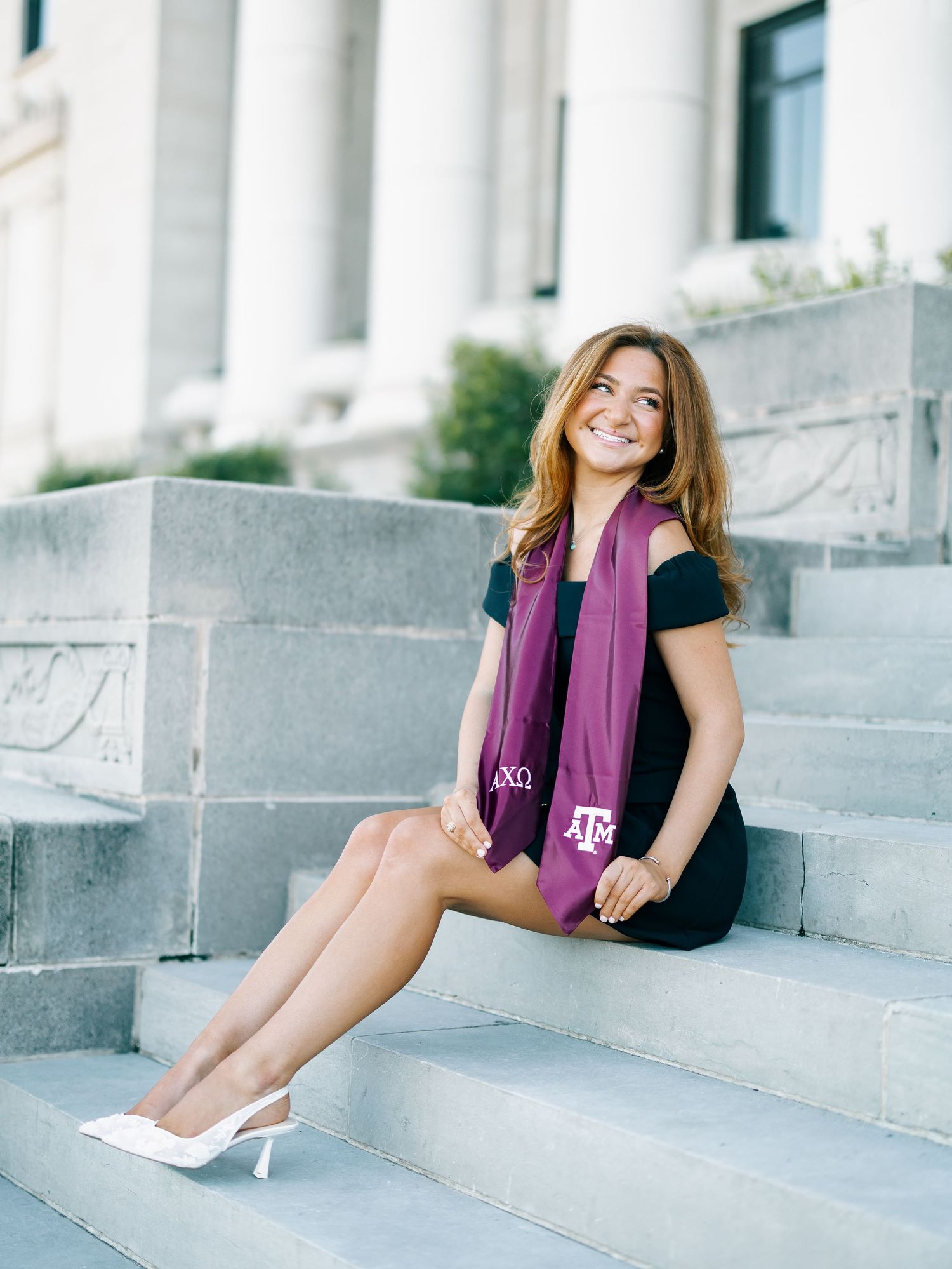 Smiling woman in black dress with Texas A&M maroon graduation stole sitting on stone steps.
