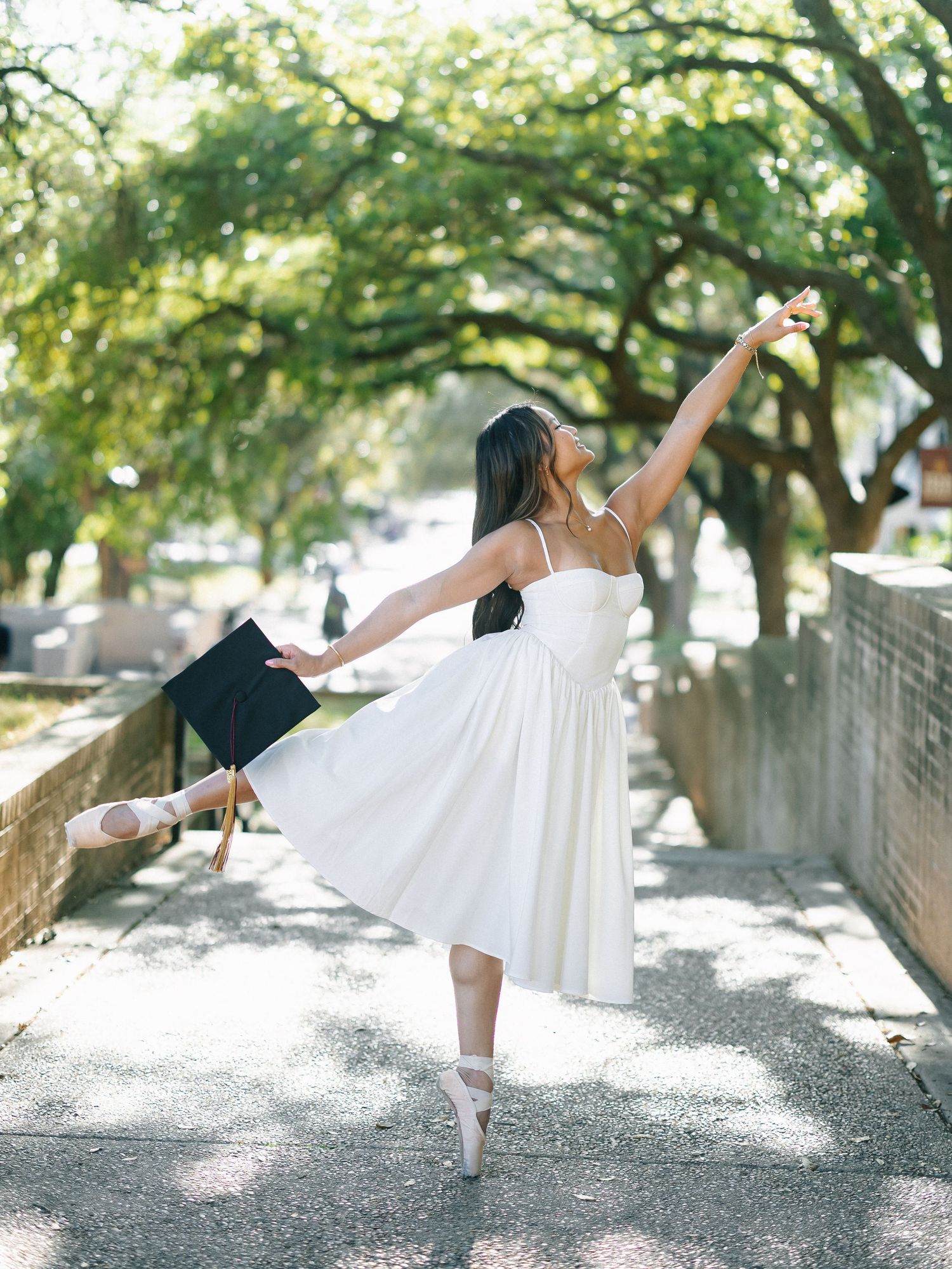 Ballerina in white dress and pointe shoes holds graduation cap while dancing on a tree-lined path.