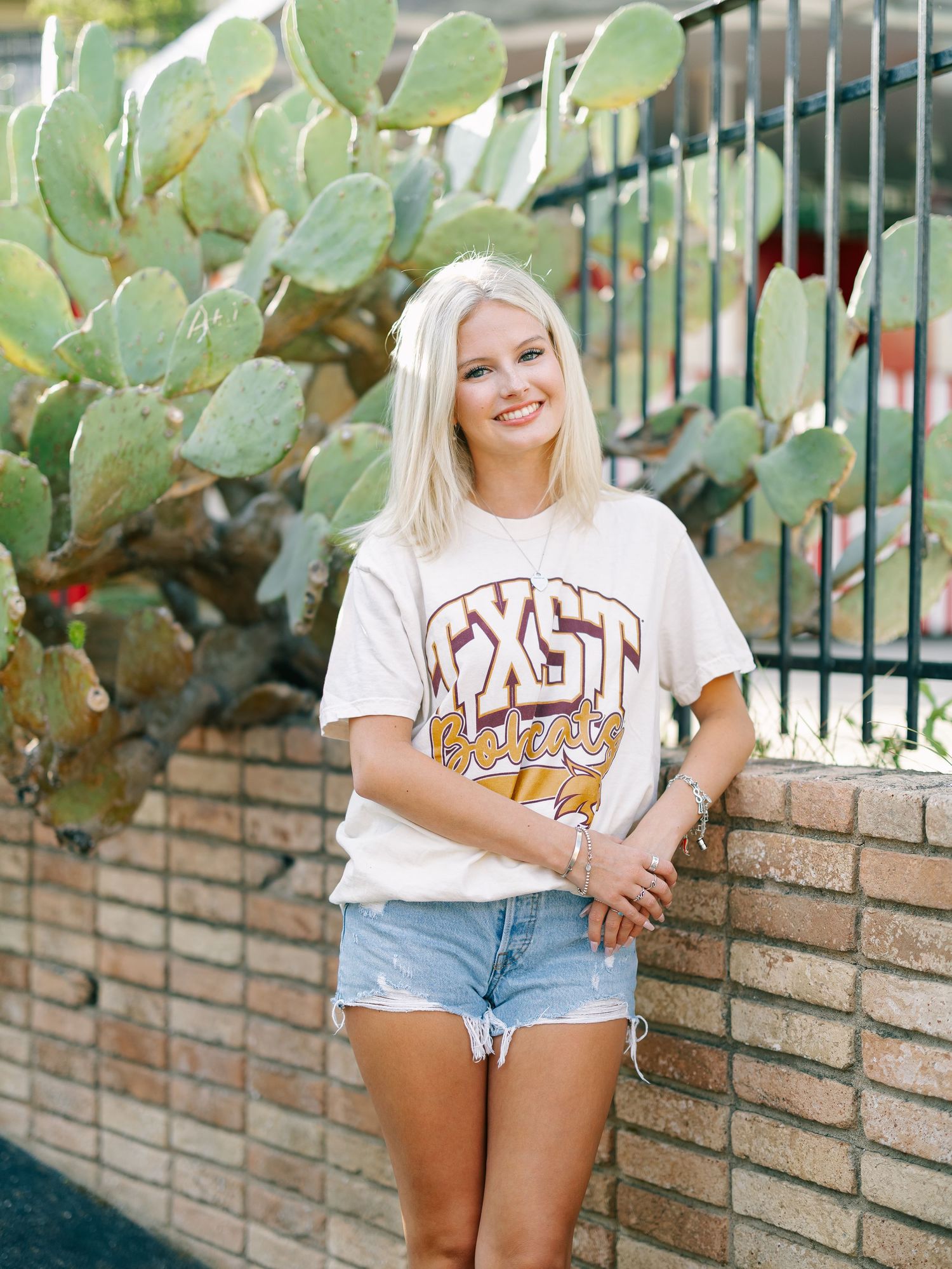 Smiling blonde woman in TXST t-shirt and denim shorts leaning against brick wall with cactus backdrop.