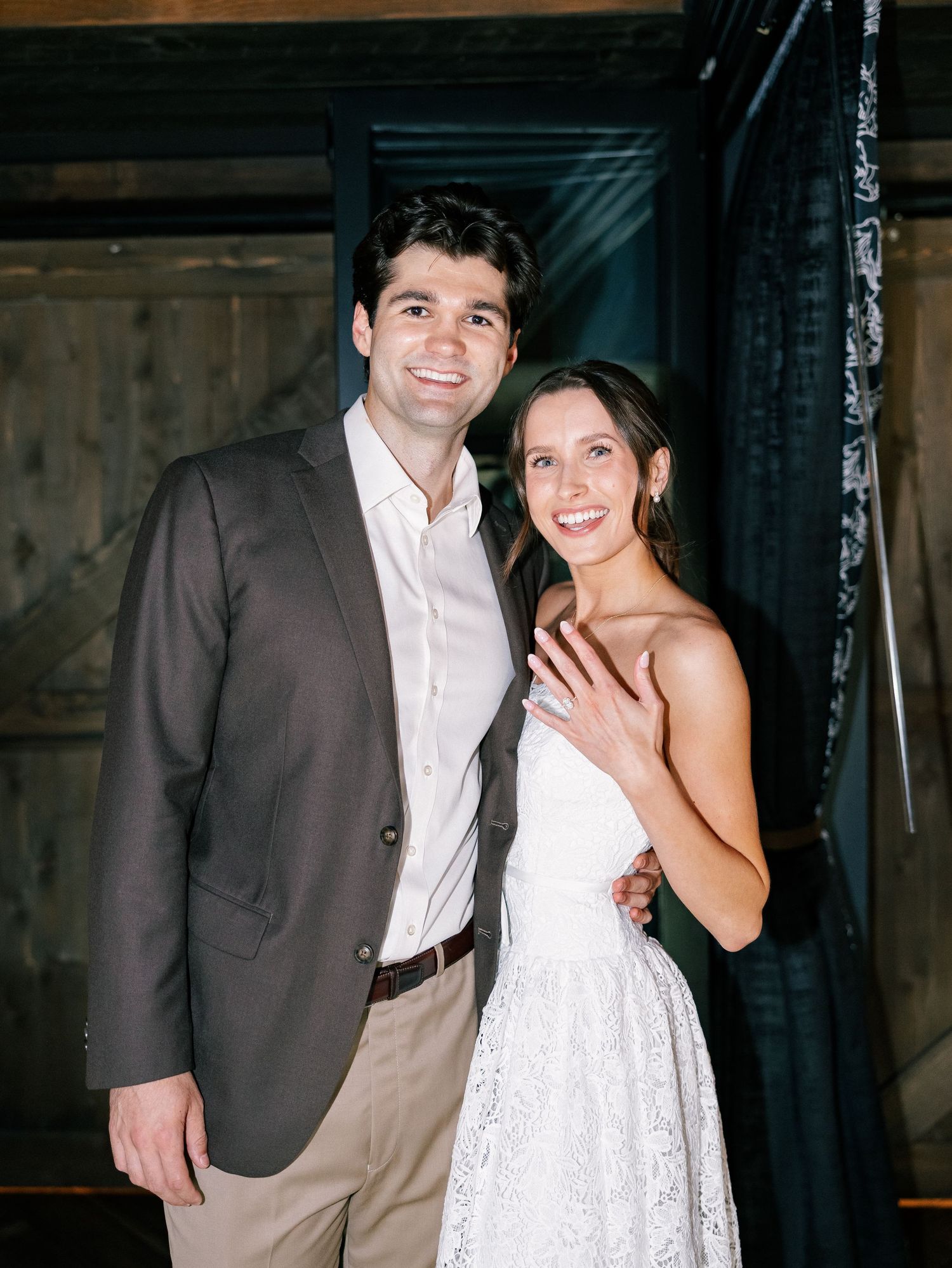 Happy couple posing together at a rustic venue, woman displaying engagement ring in white lace dress.