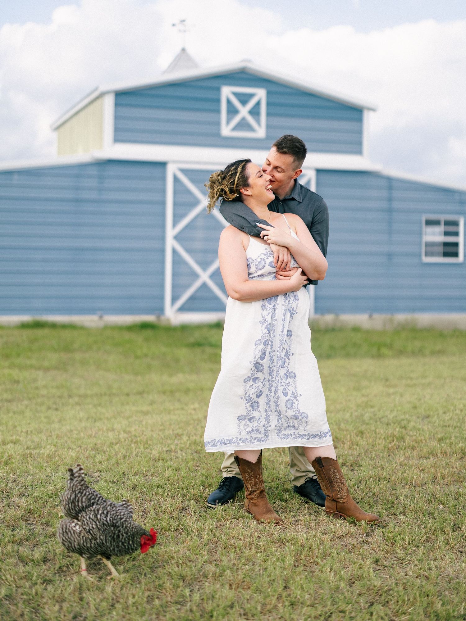 Couple embracing in front of a blue barn on a farm with a chicken nearby.
