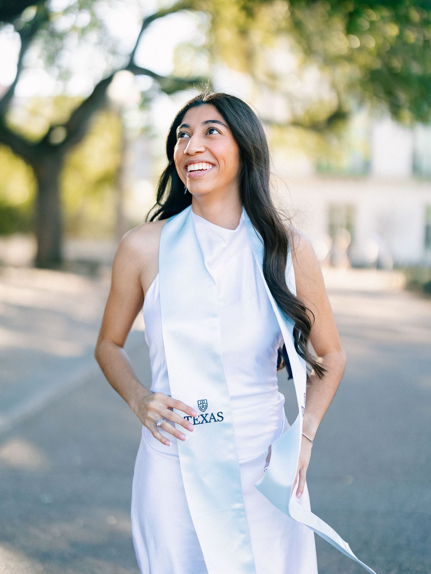 Smiling female graduate wearing white dress and Texas graduation stole poses outdoors on sunny campus.
