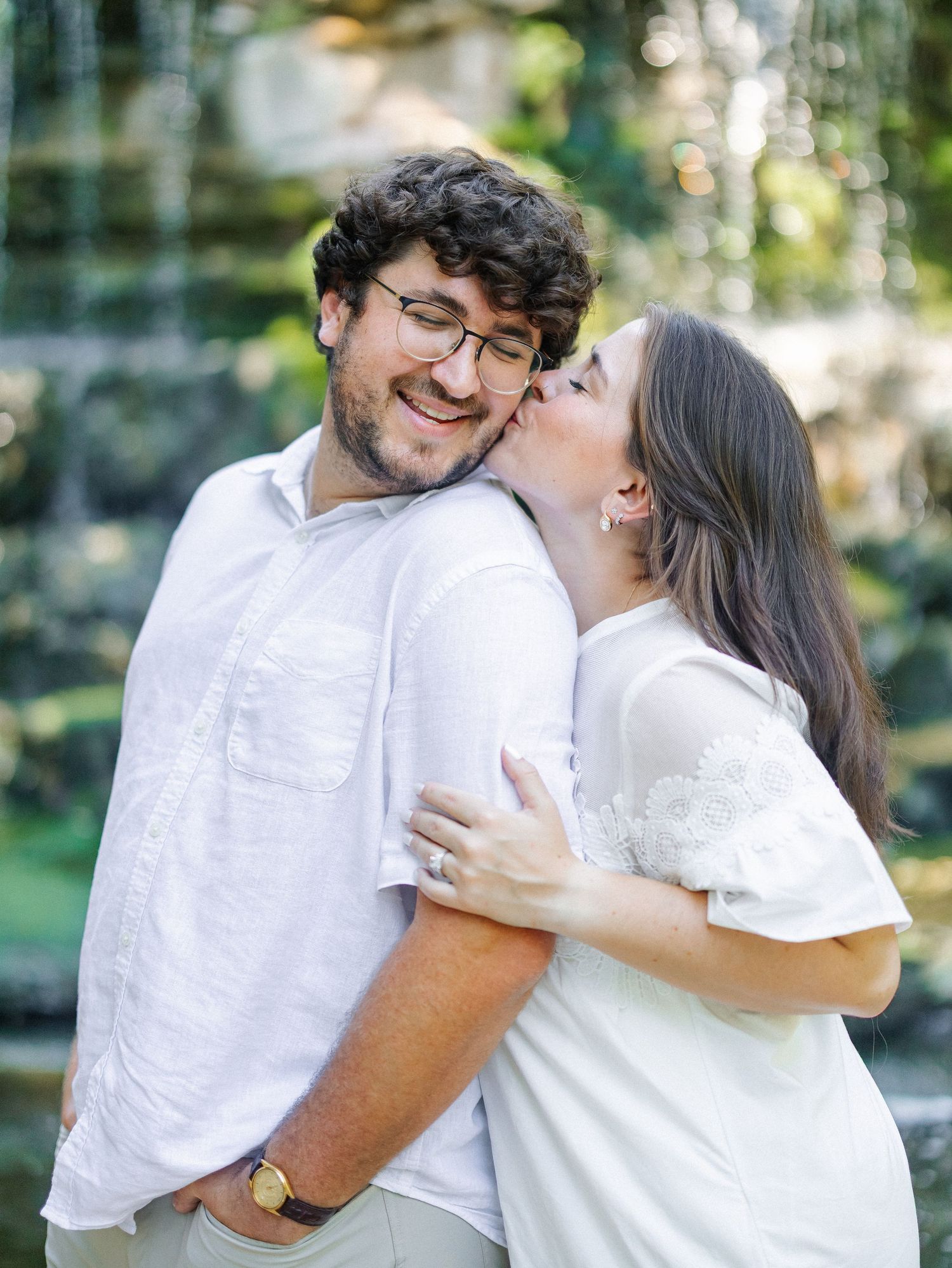 A smiling man in glasses and white shirt receives a kiss on the cheek from a woman in a white dress outdoors.