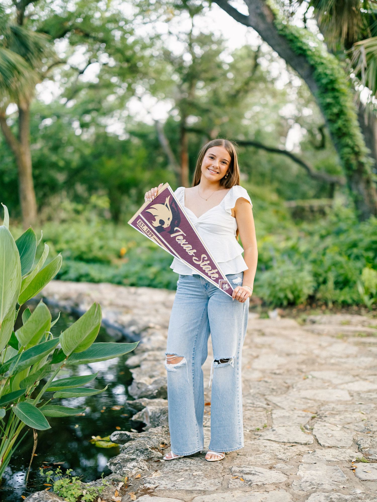 Young woman smiling and holding a Texas State University pennant sign outdoors in a lush garden setting.