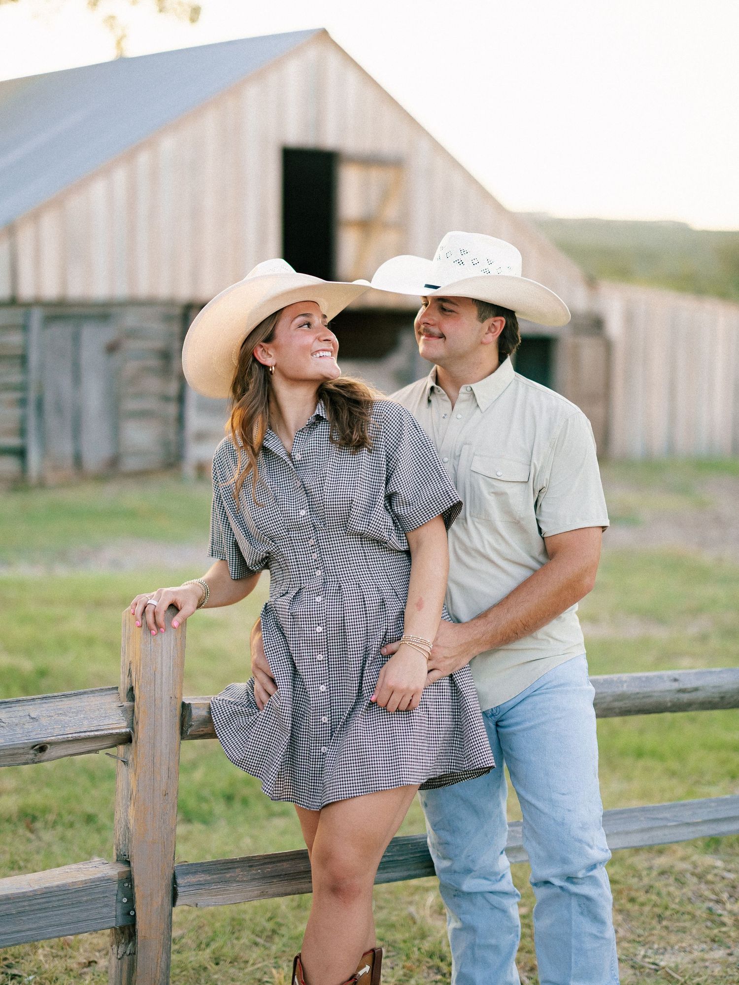 Couple in cowboy hats smiling by a wooden fence with a rustic barn backdrop during golden hour.