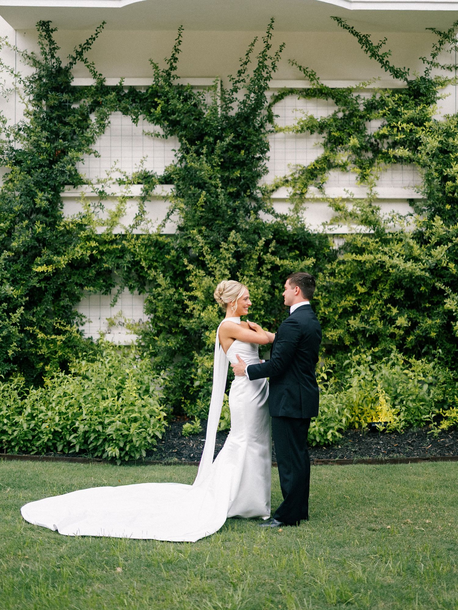 Bride and groom share a romantic moment on a green lawn with ivy-covered white trellis backdrop.