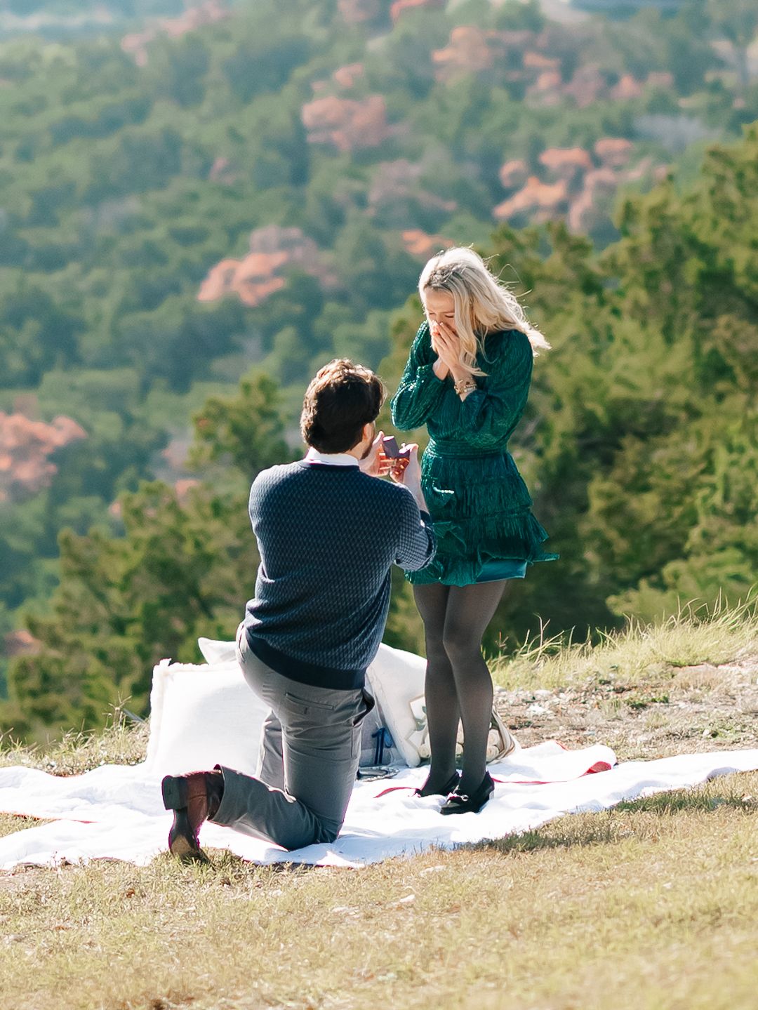 Man proposing on one knee to surprised woman on scenic hilltop overlook.