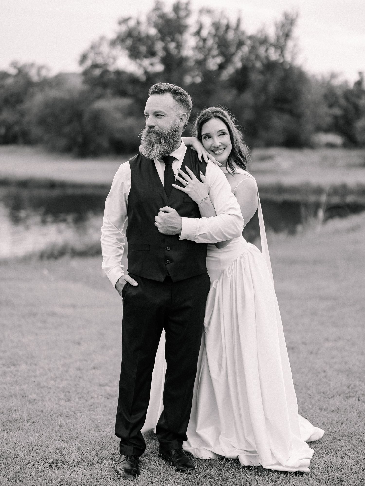 Black and white photo of a bride in a flowing gown embracing a bearded groom in a vest by a pond.