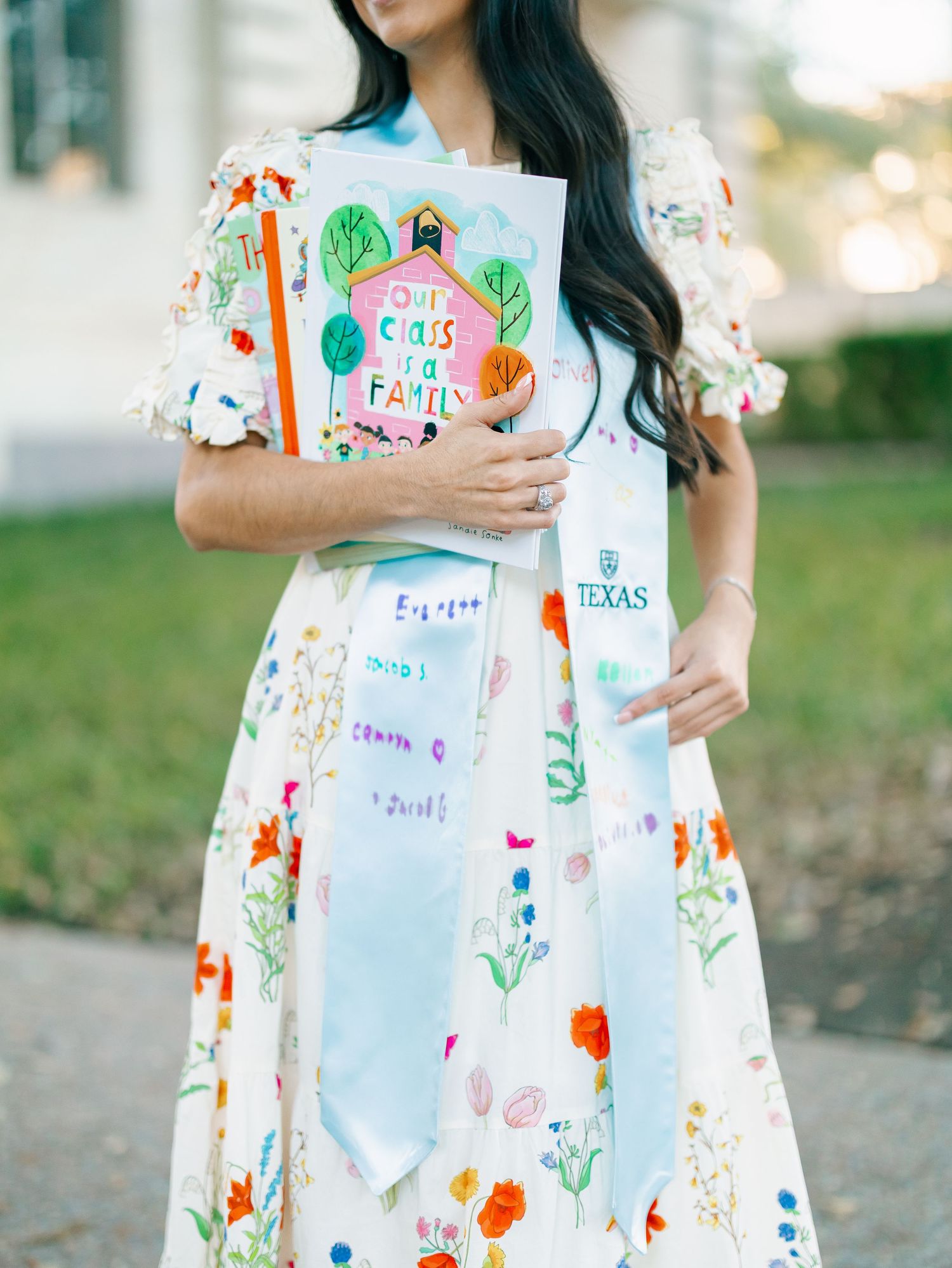 Woman in floral dress holding a colorful book and Texas graduation sash at a graduation celebration.