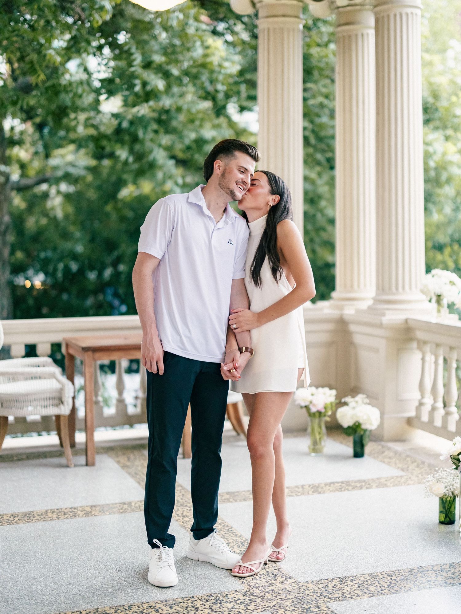Couple sharing a kiss on an elegant columned porch with white floral arrangements nearby.
