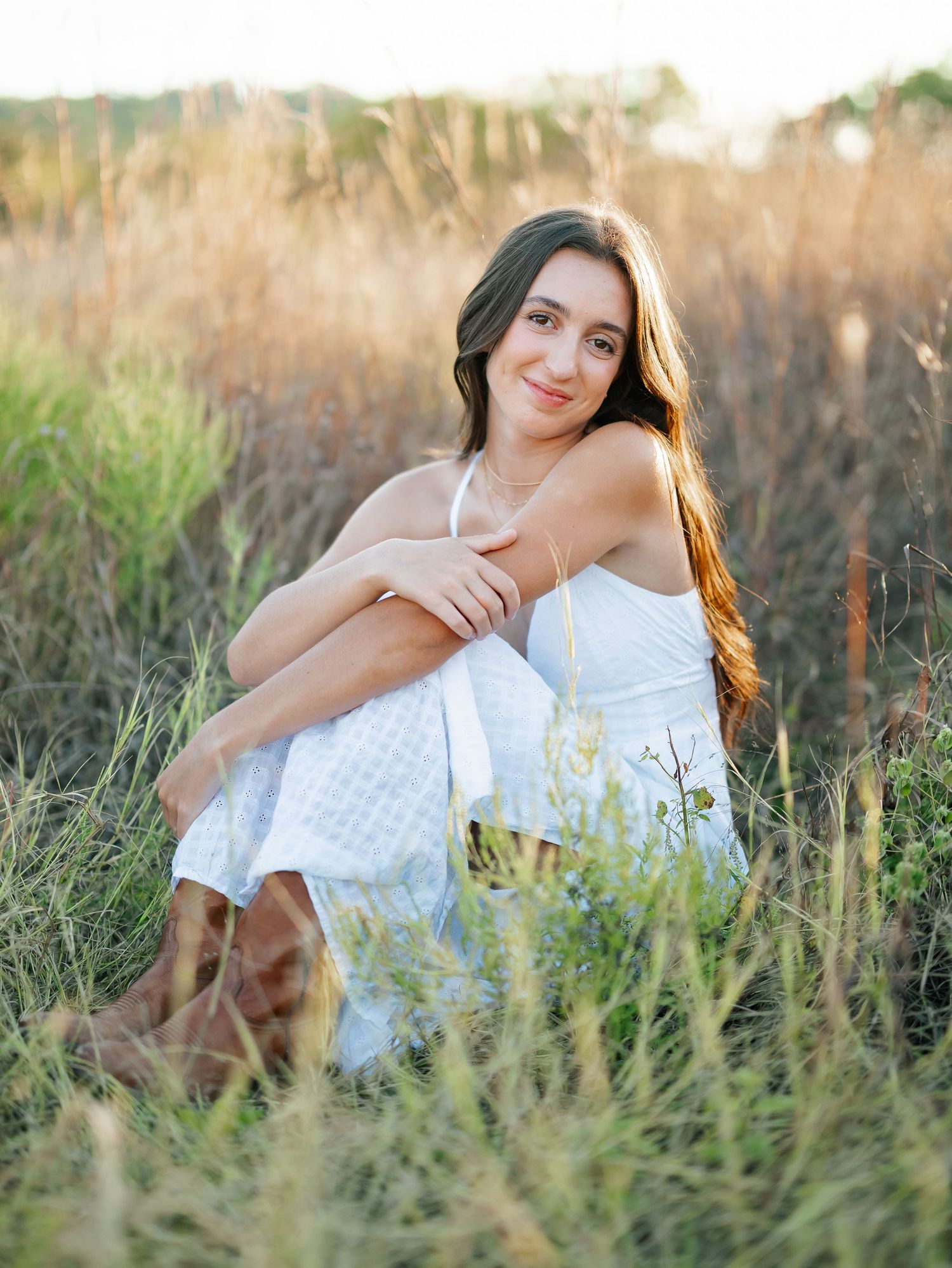 Young woman in white dress and brown boots sitting in golden field grass during sunset portrait session.