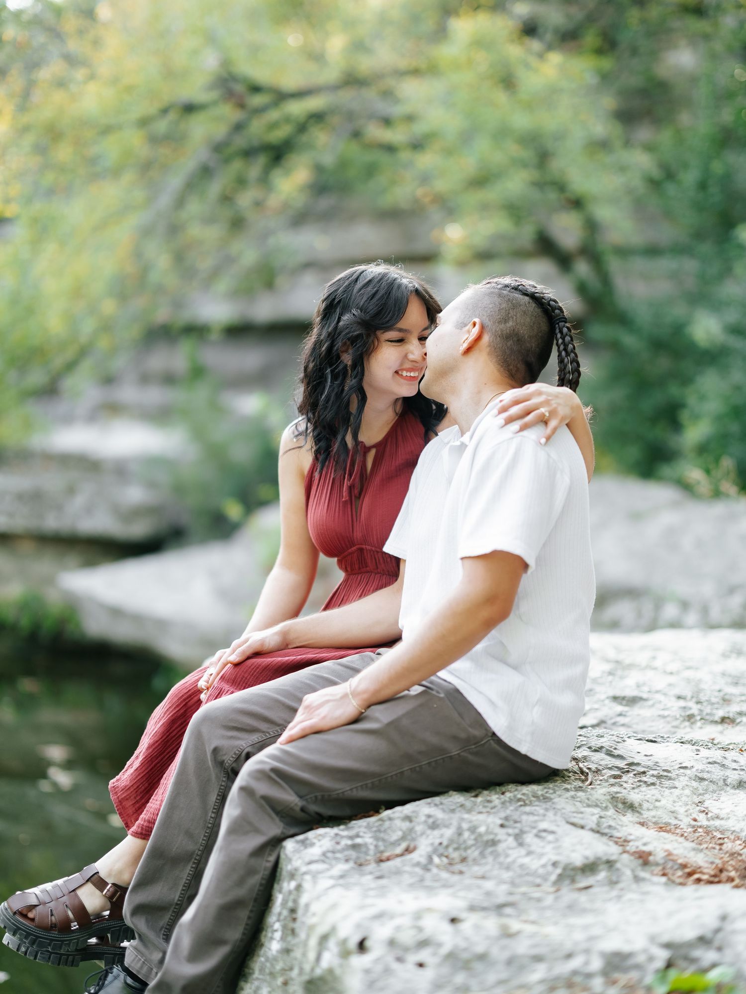 Couple sitting on a rock by a creek, sharing a tender moment surrounded by green trees.