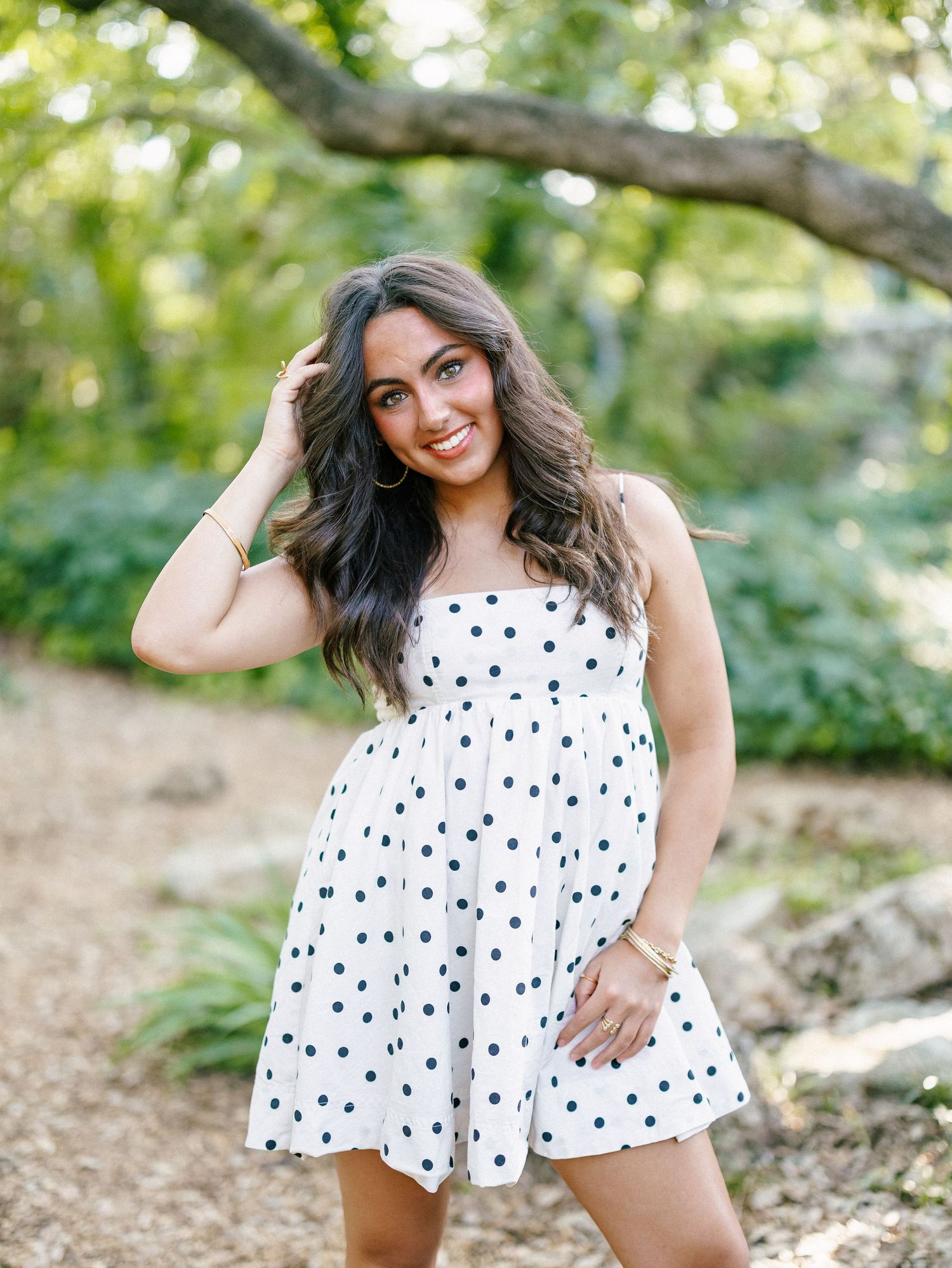 A smiling young woman in a white polka dot romper stands among trees with her hand in her hair.