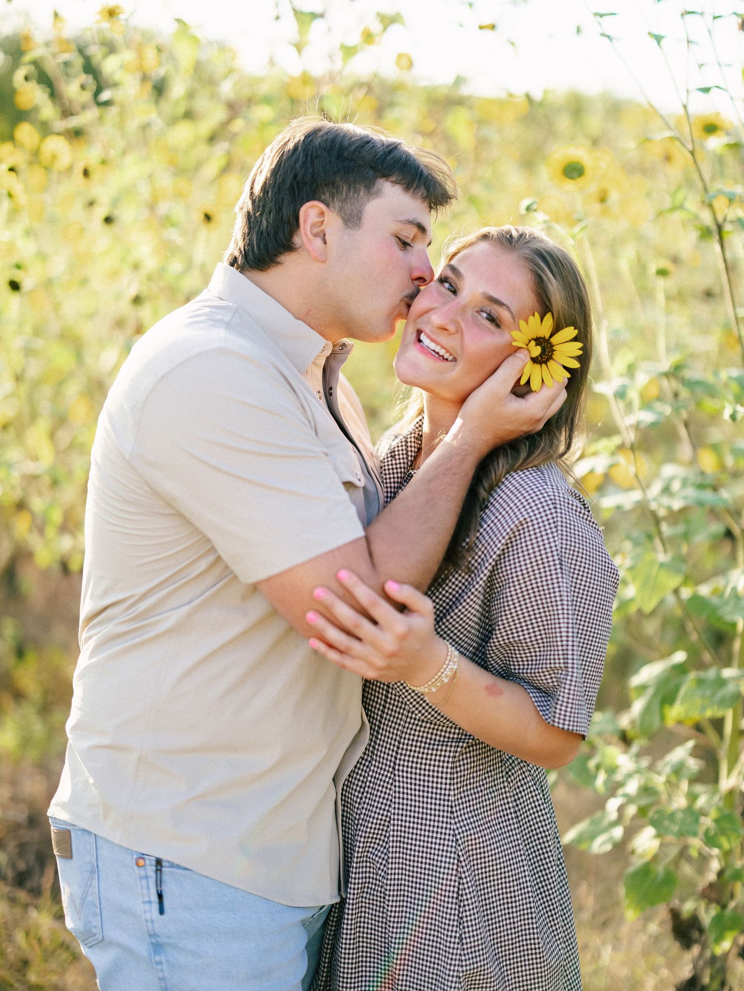 Couple embracing in sunflower field, man kissing woman's cheek as she smiles holding a yellow sunflower.