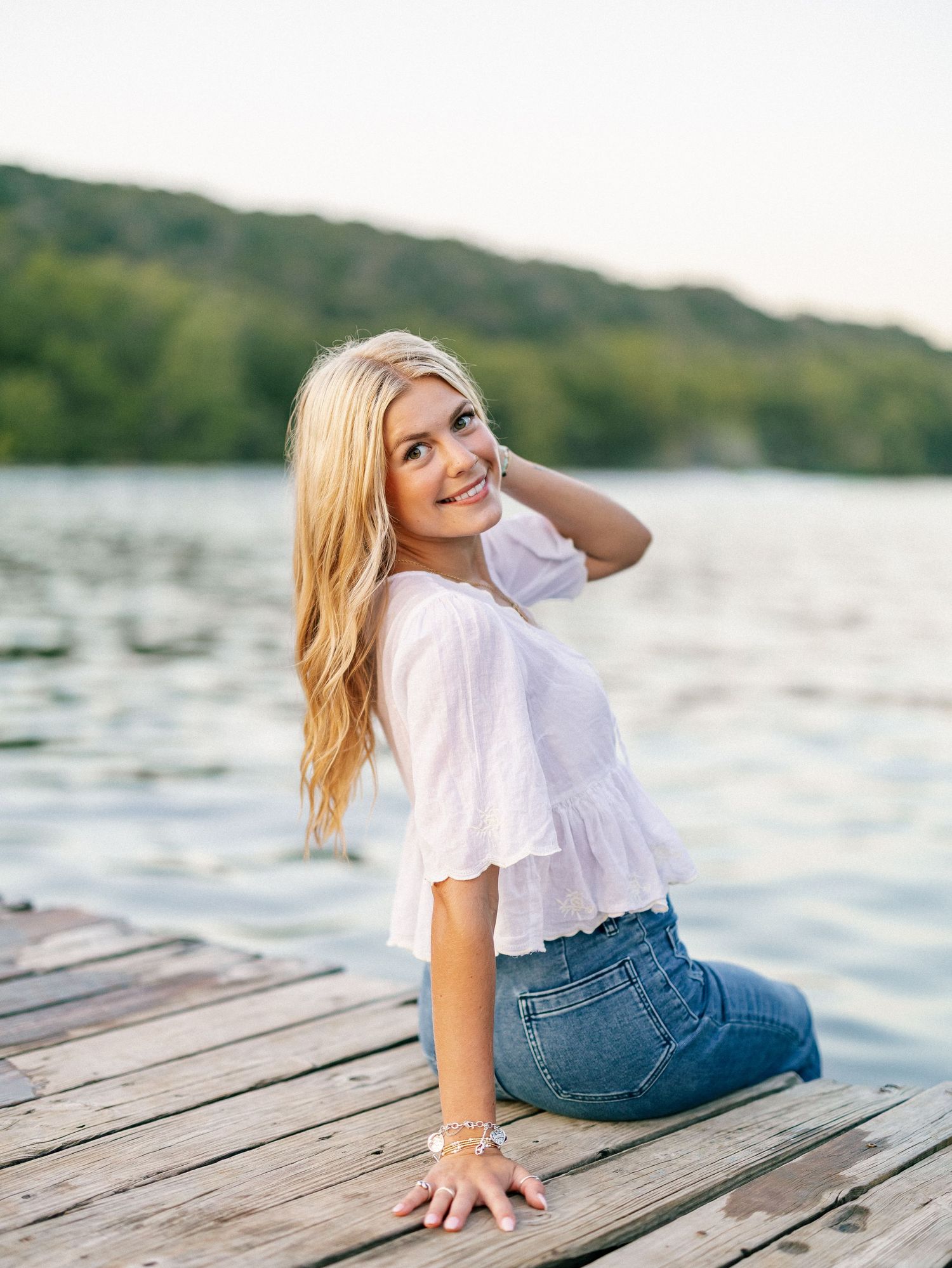 Young blonde woman sitting on a wooden dock by a lake, wearing a white top and jeans, smiling over her shoulder.