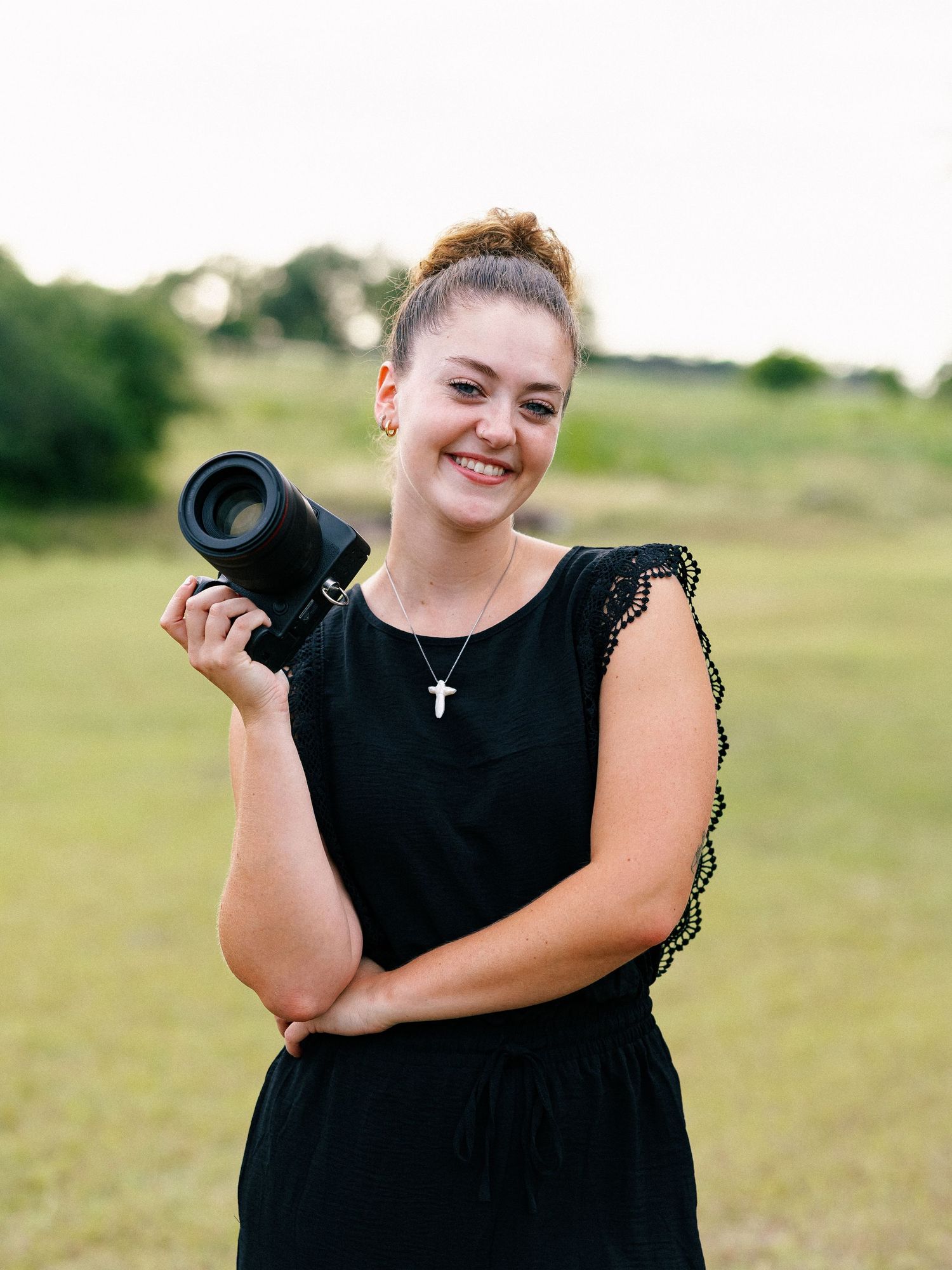 Smiling young woman in black dress holding a camera outdoors in a green field, wearing a cross necklace.