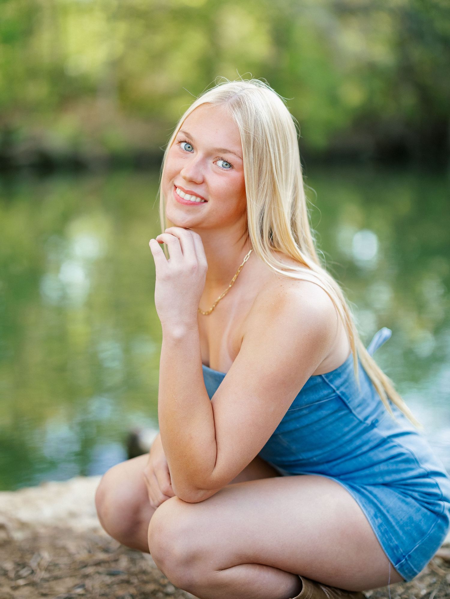 Young blonde woman smiling outdoors near a reflective pond, wearing a blue denim romper in a natural setting.