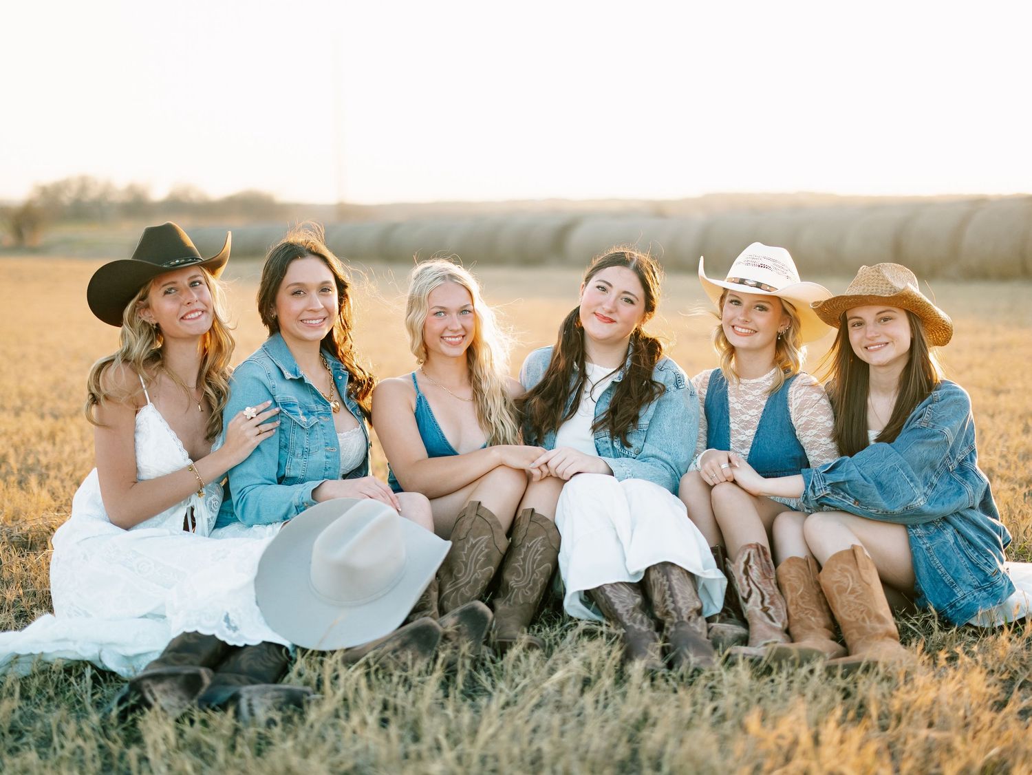 Six young women in cowboy hats and denim sit together smiling in a golden field at sunset.