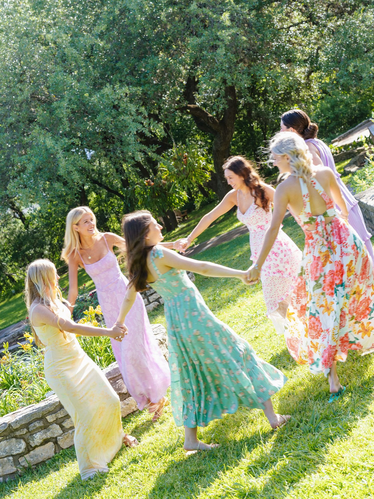 Six women in colorful floral maxi dresses hold hands dancing in a circle on a sunny green lawn.