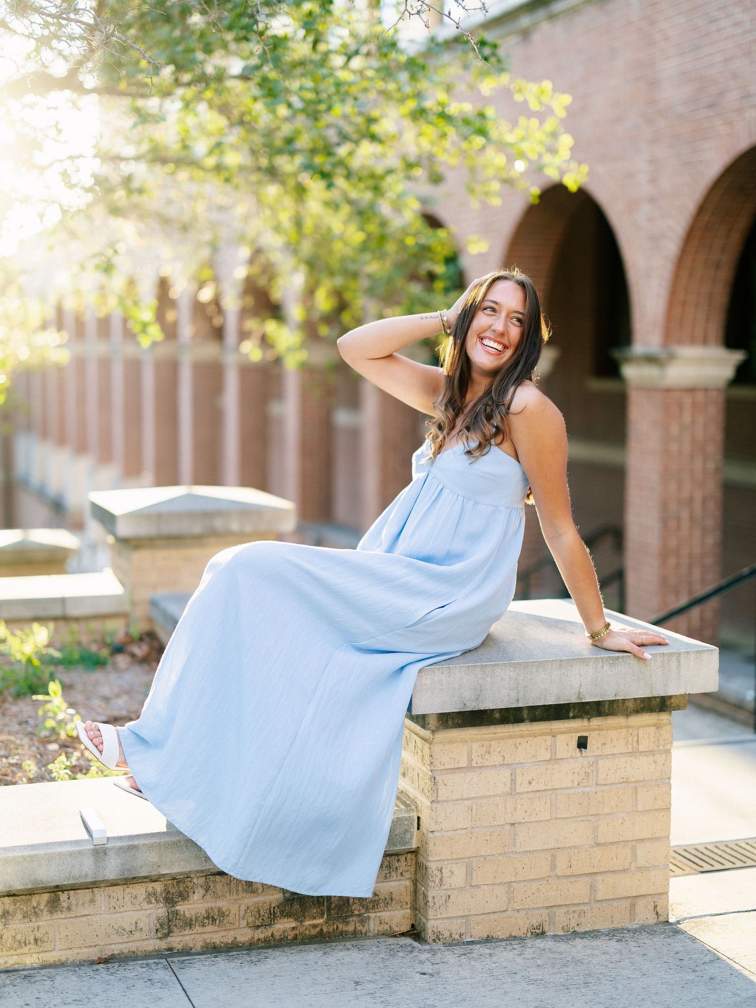 Young woman in light blue maxi dress sitting on brick wall outdoors, smiling, with arched building in background.