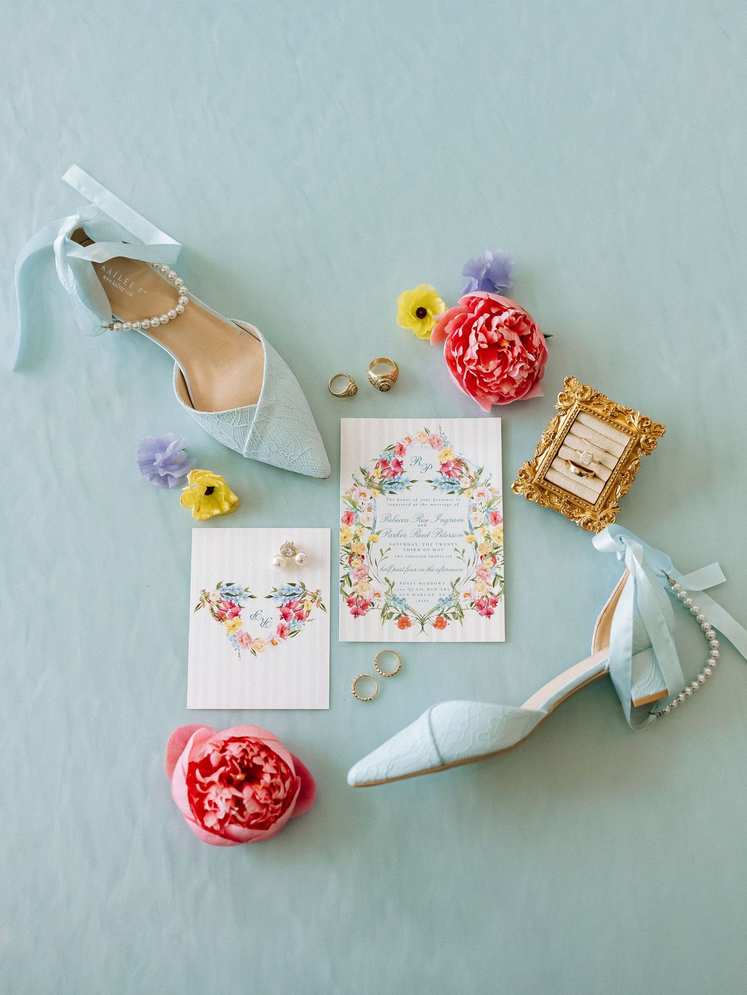 Flat lay of light blue wedding heels, floral invitations, rings, and pink peonies on blue background.