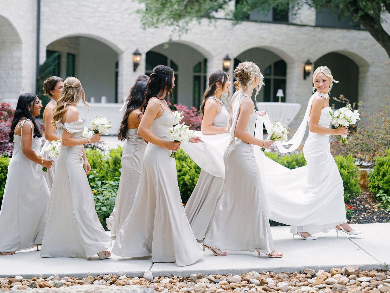 Bridesmaids in elegant gray gowns walk with white bouquets outside a stone venue on a sunny wedding day.