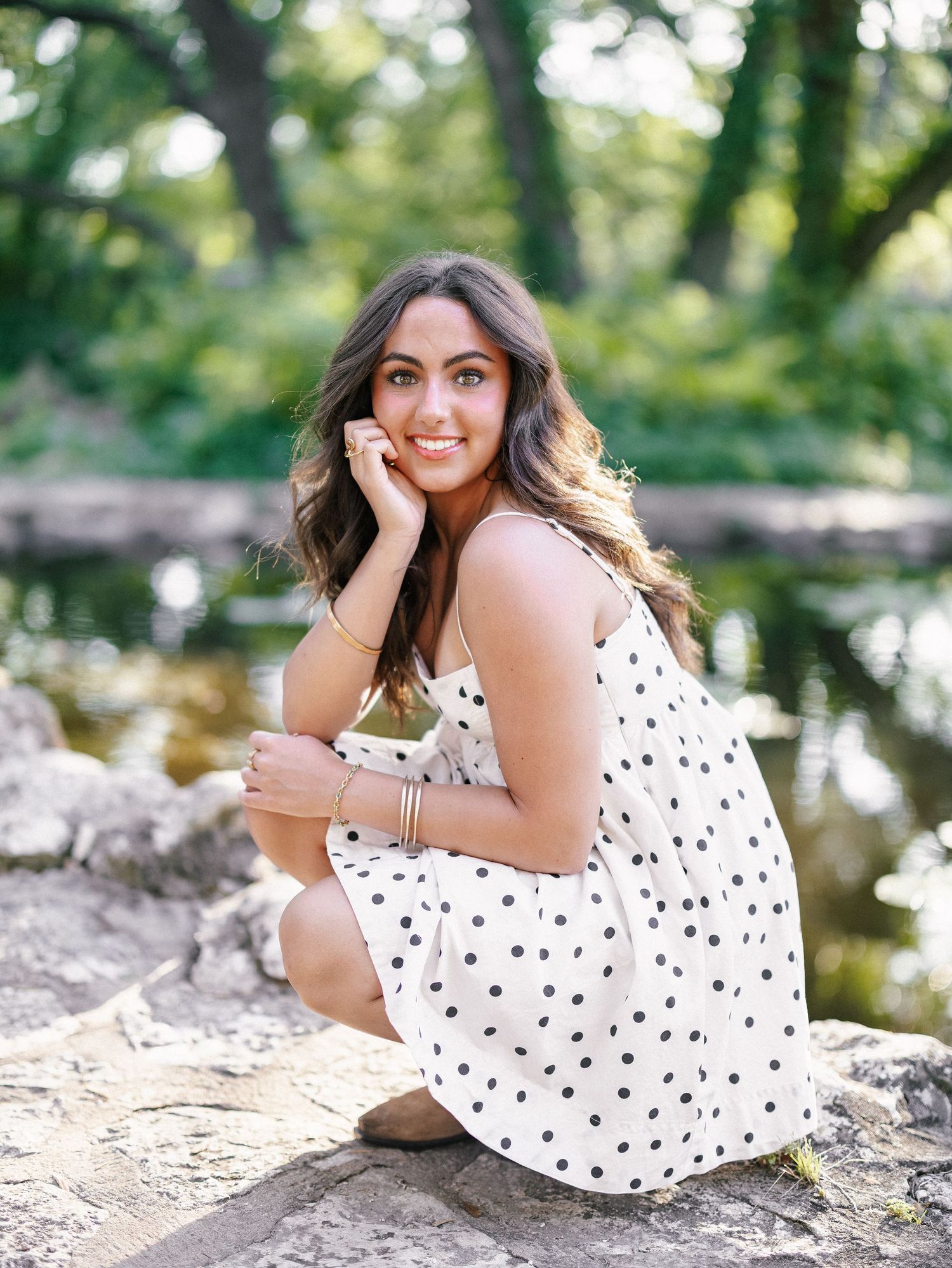 Young woman in white polka dot dress poses on rocks near a pond in a lush green outdoor setting.