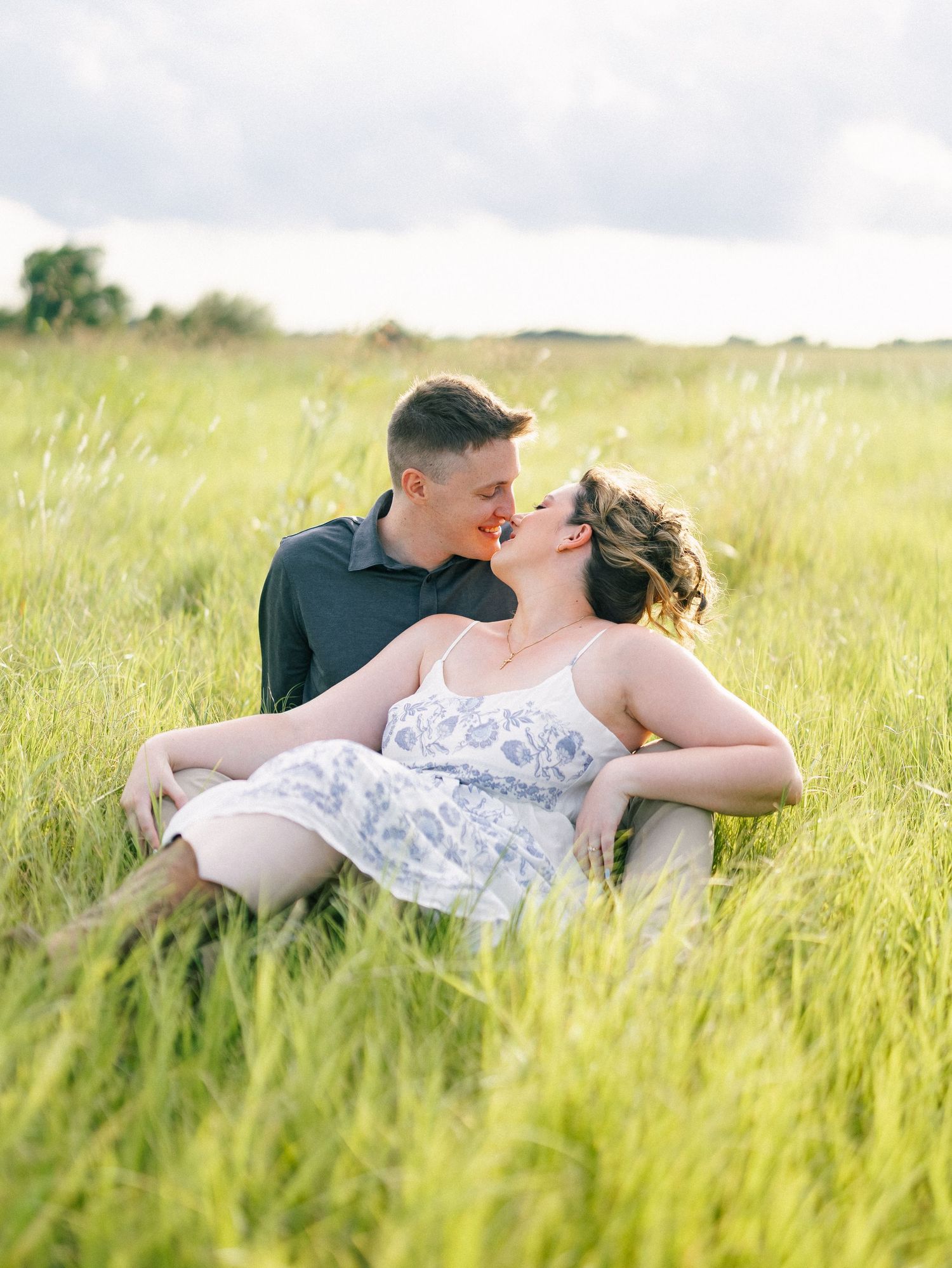Couple sharing a romantic kiss while sitting in a lush green field during an outdoor engagement photo session.