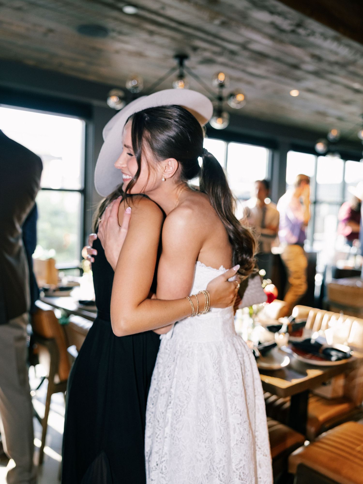Two women embracing at a wedding reception, one in a white lace dress and hat, the other in a black dress.
