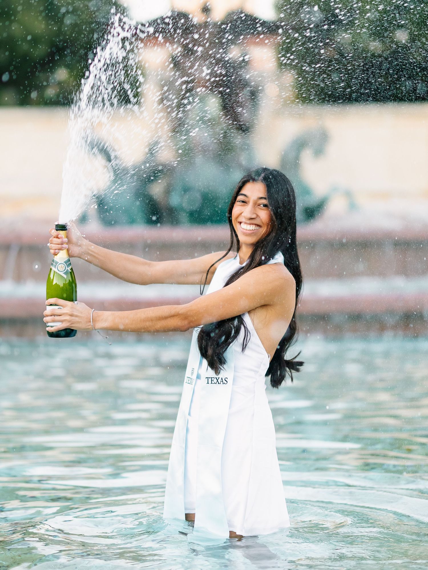 Smiling woman in white Texas dress pops champagne bottle while standing in outdoor fountain.