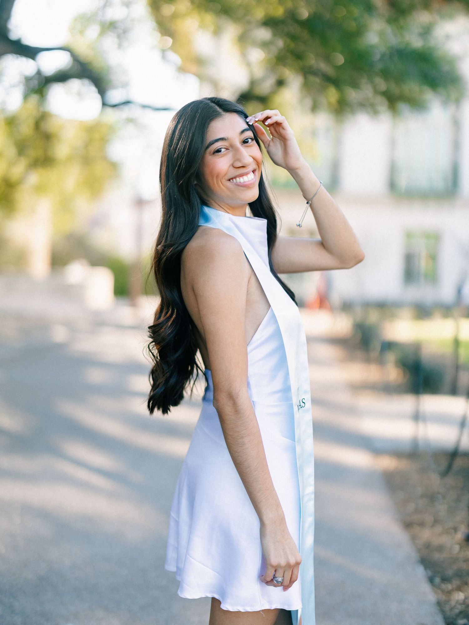 Smiling young woman in white graduation dress posing outdoors on a sunny day with long dark wavy hair.