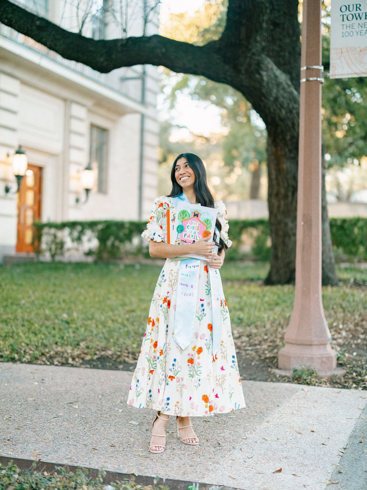 Young woman in floral white midi dress holding diploma, smiling outdoors near decorative post on graduation day.