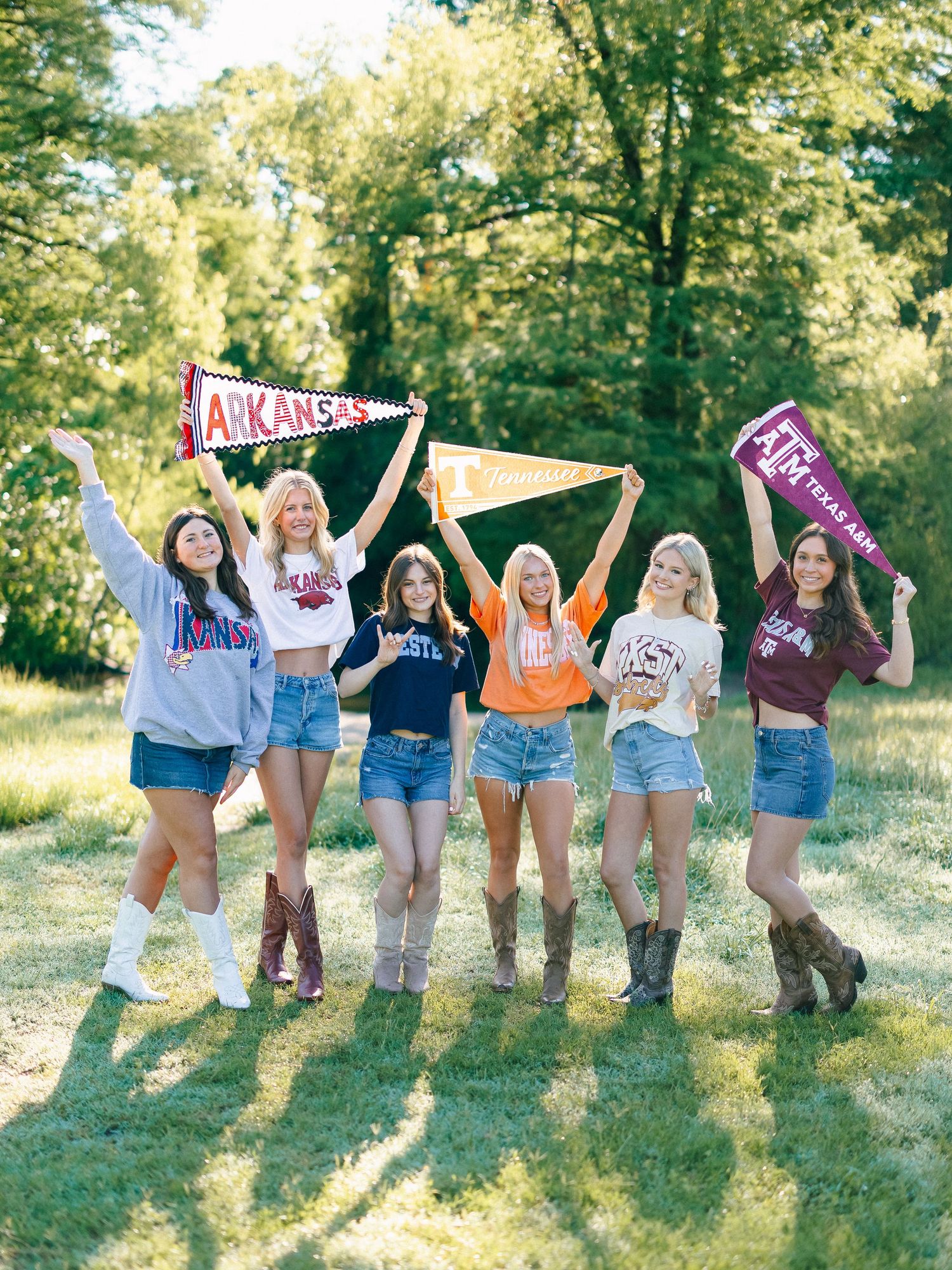 Six young women holding college pennants including Arkansas and Texas A&M, wearing school shirts and denim shorts outdoors.