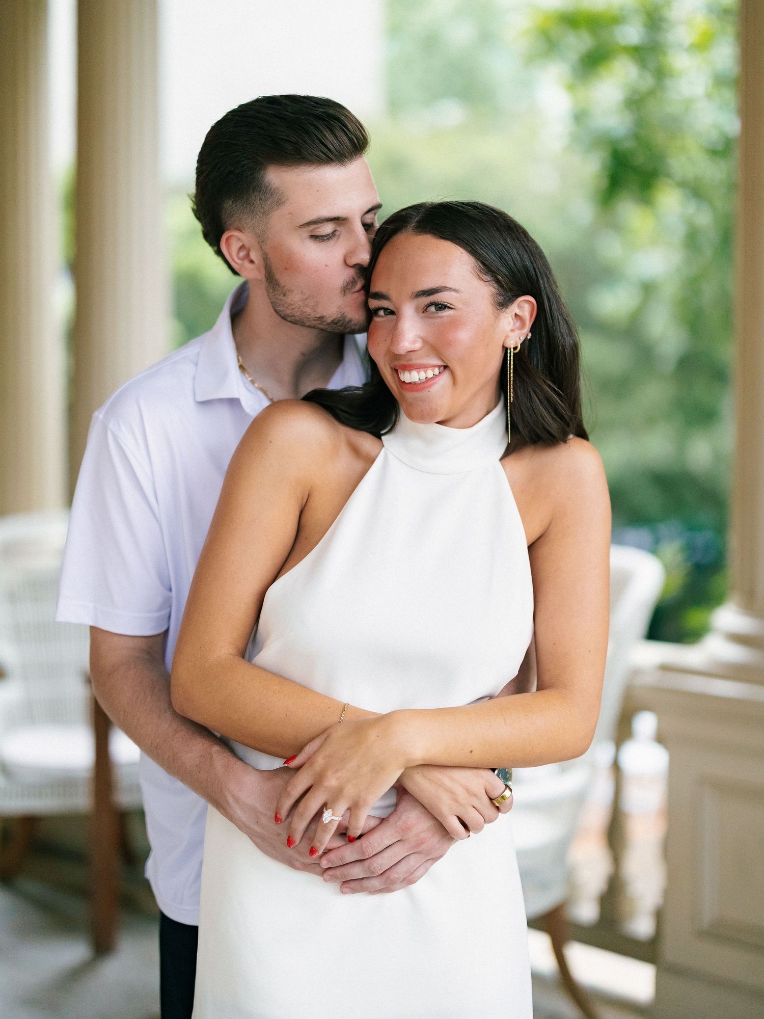 Couple embracing on a porch, woman in white halter dress smiling as man kisses her cheek during engagement photo session.