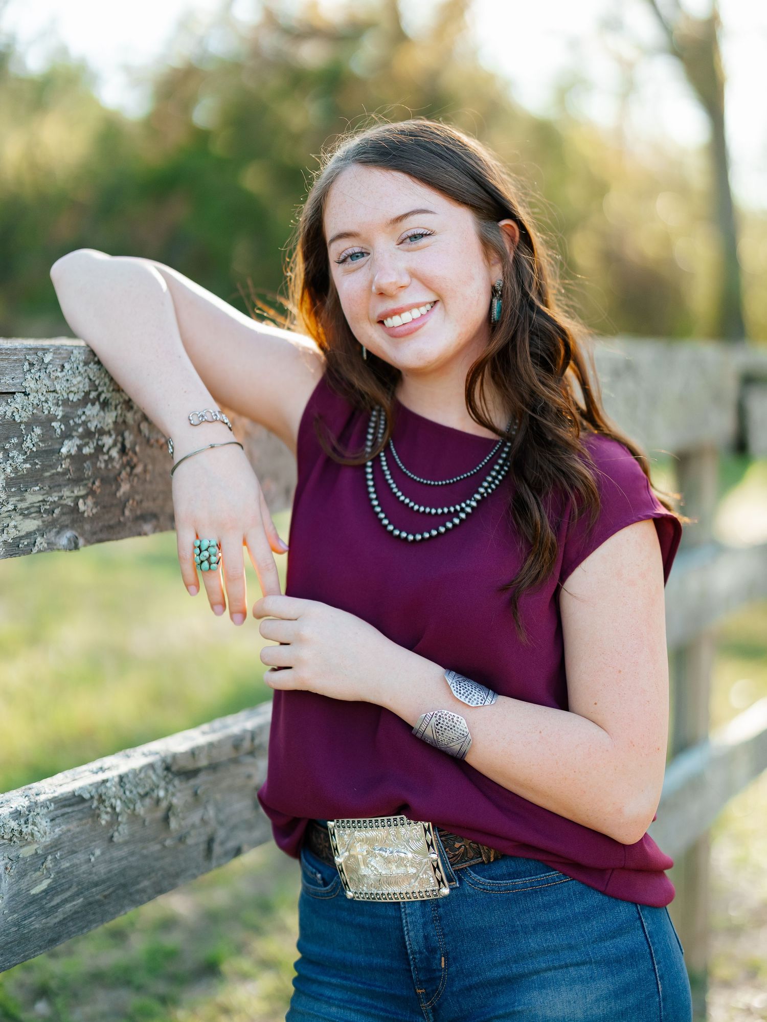 Smiling young woman in maroon top, turquoise jewelry, and decorative belt poses by wooden fence outdoors.