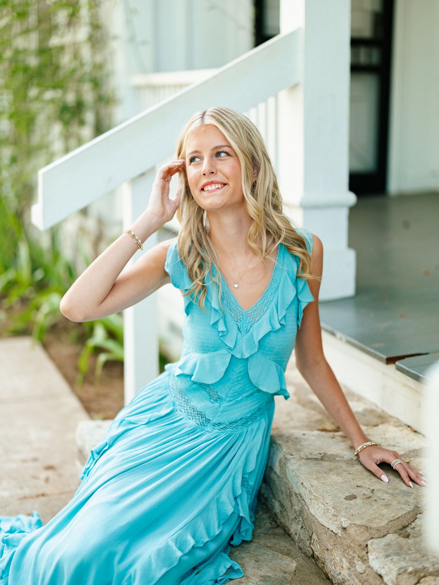 Blonde woman in turquoise ruffle maxi dress sitting on stone steps outdoors, smiling gracefully.