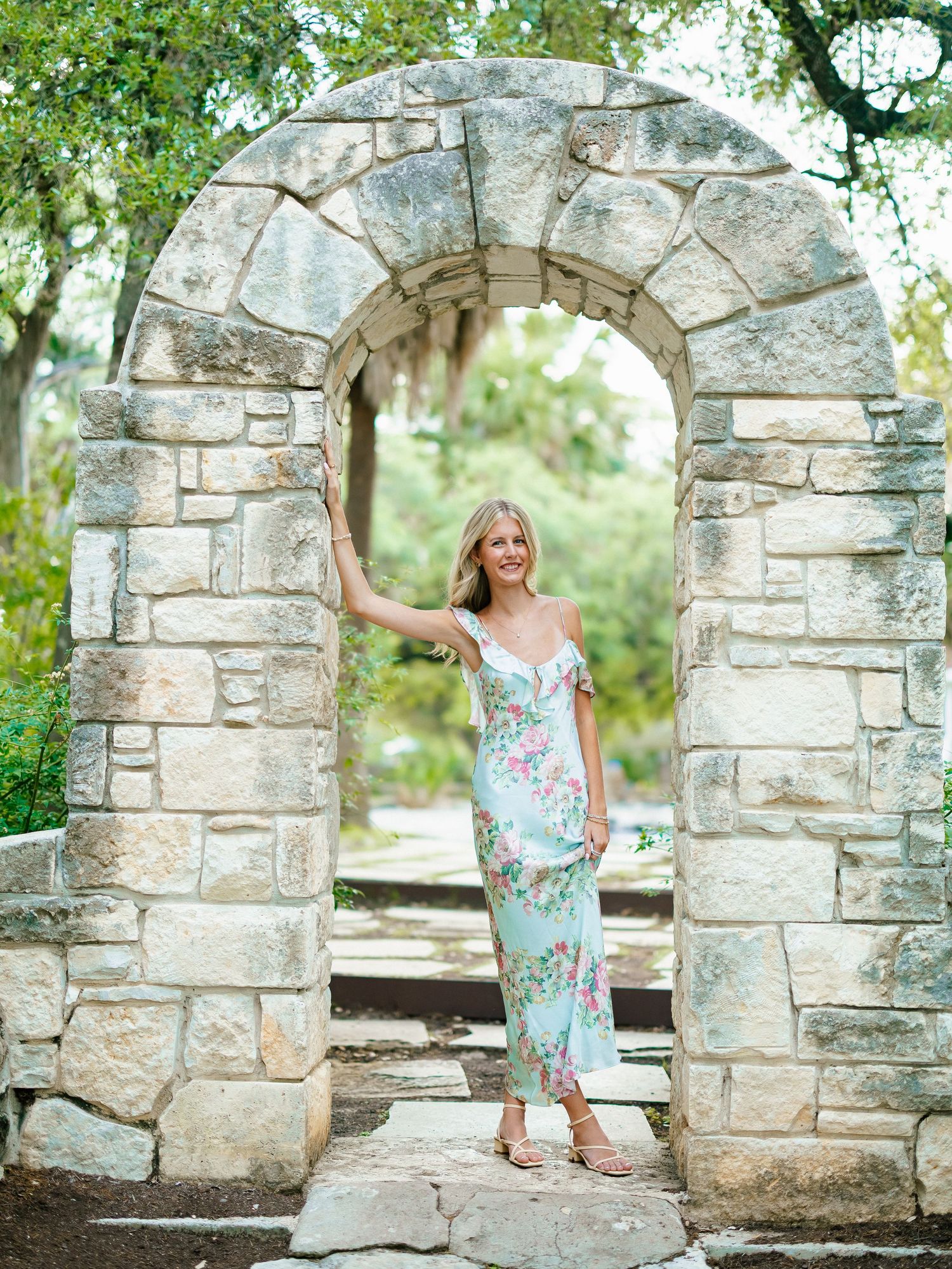 Elegant woman in floral midi dress posing under a rustic stone arch in a garden.