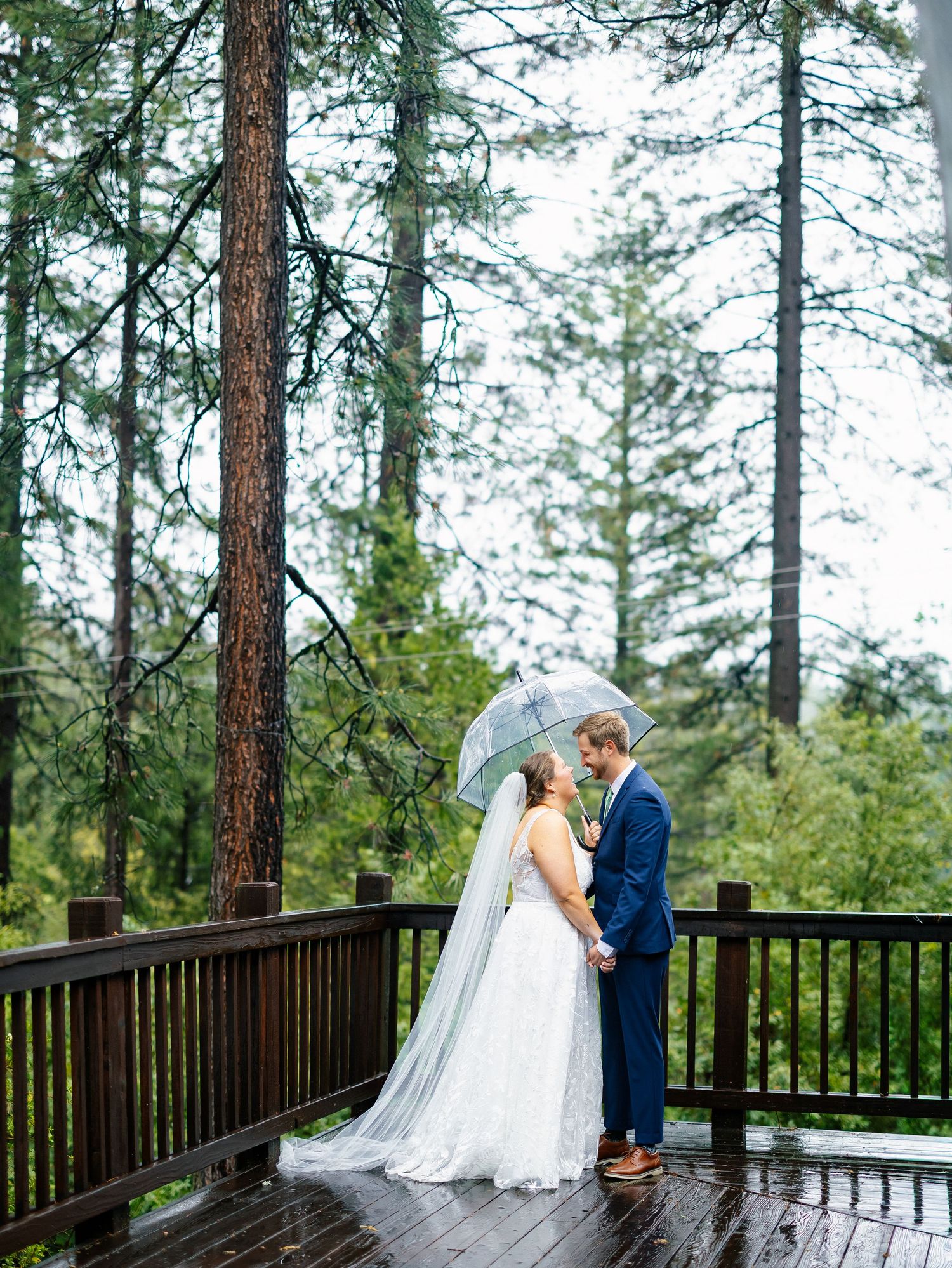 Bride and groom share a romantic moment under a clear umbrella on a rainy forest deck.
