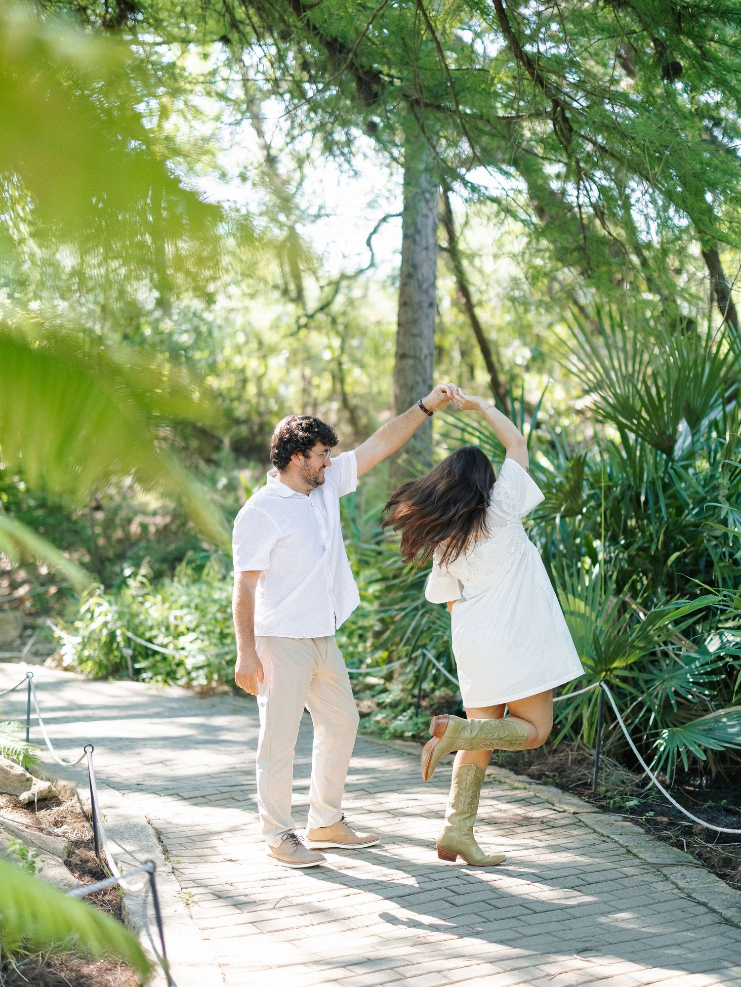 A couple dances joyfully on a garden path surrounded by lush greenery, dressed in white outfits and tan boots.