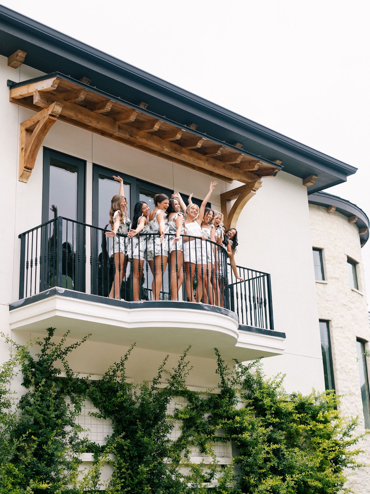 Bridal party cheers from a balcony of a modern white building draped in ivy.