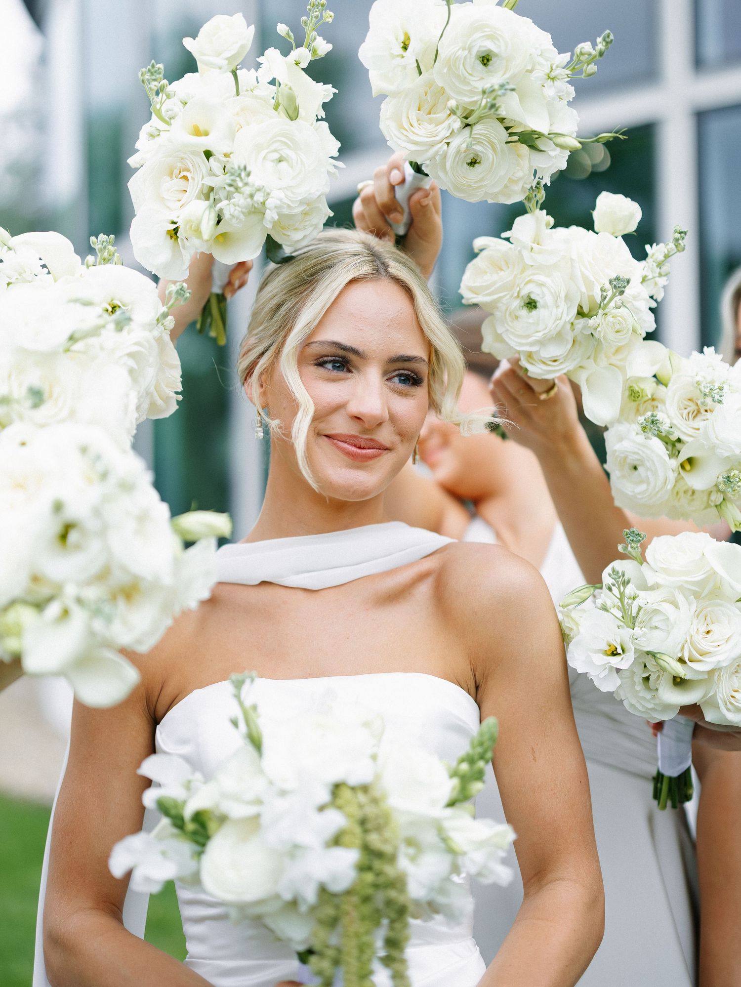 Bride surrounded by bridesmaids holding white floral bouquets featuring ranunculus and roses at a wedding.