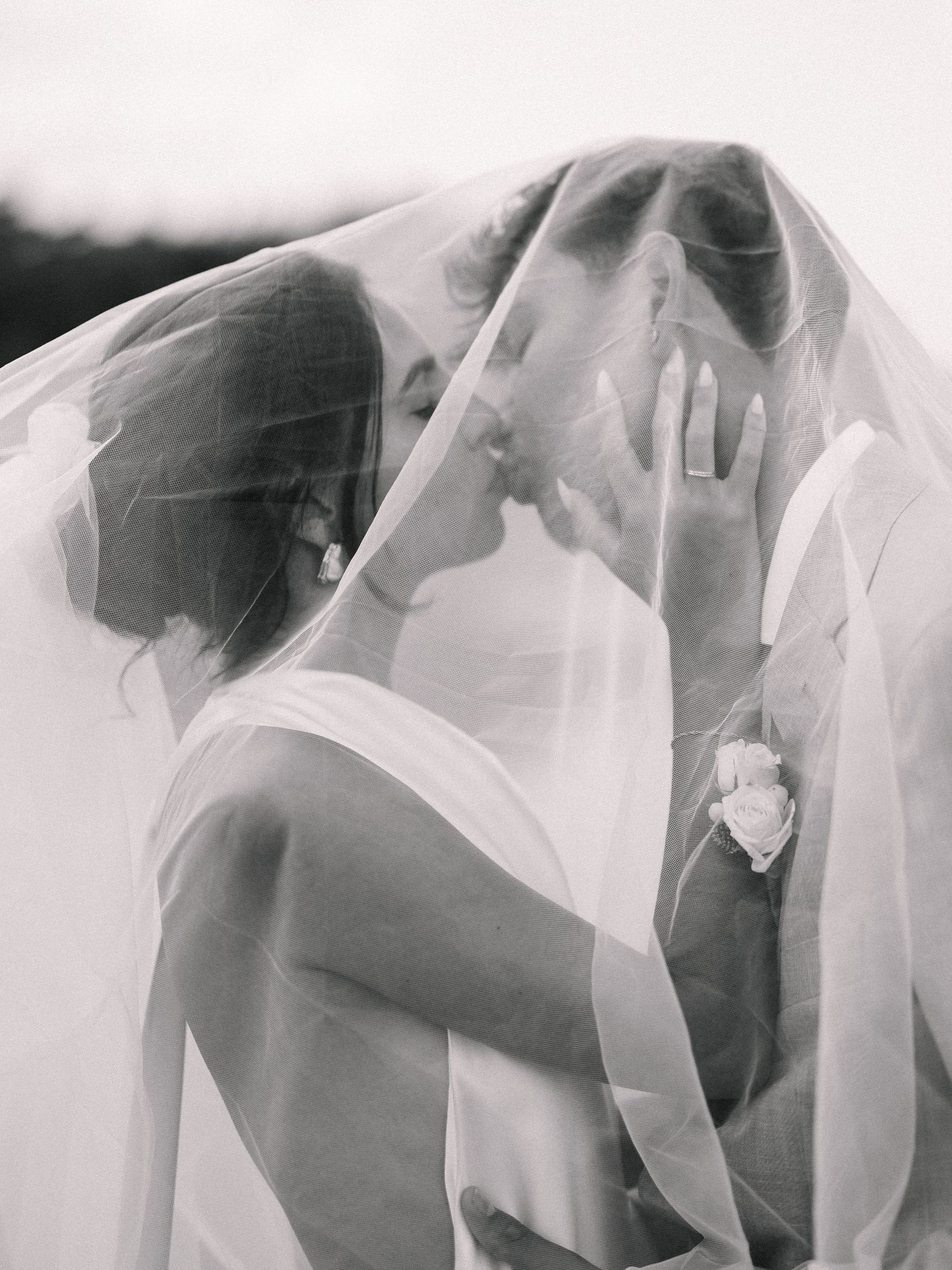 Black and white photo of bride and groom sharing a romantic kiss under a flowing wedding veil.